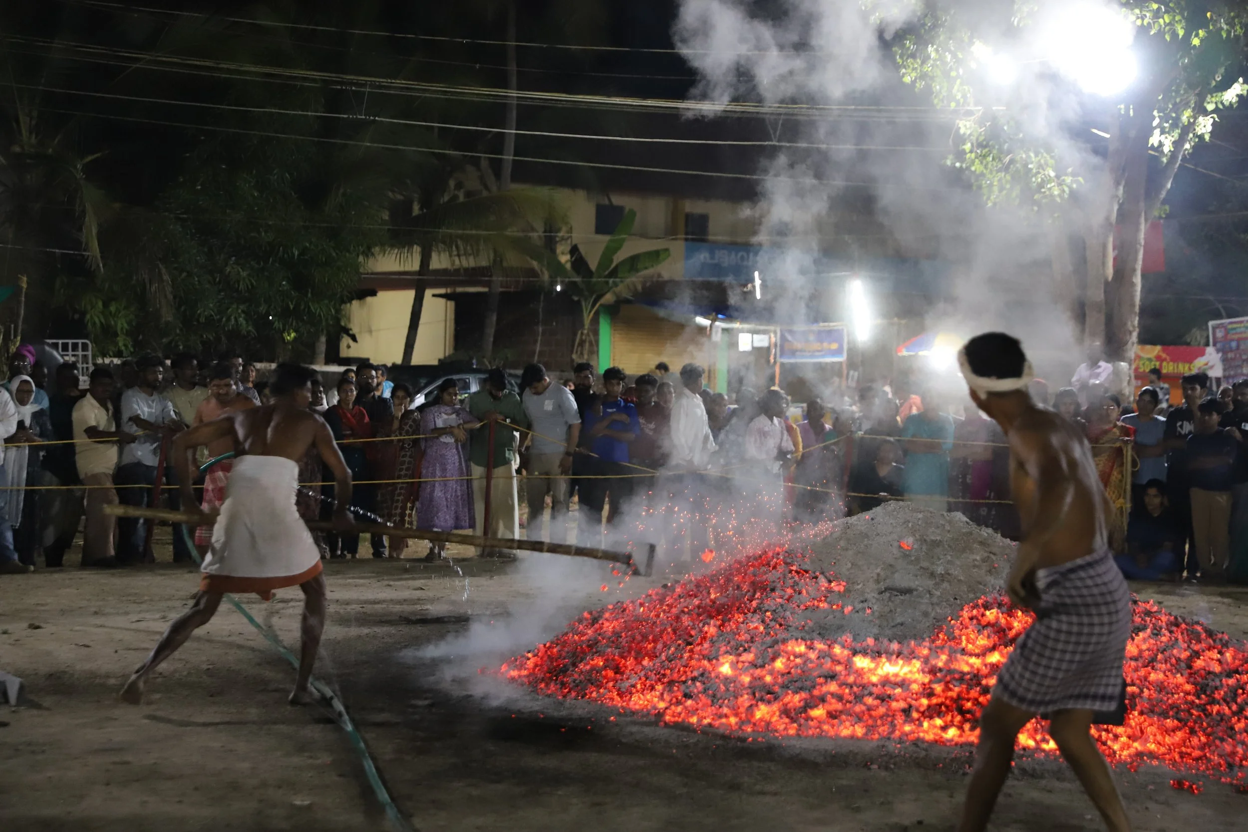 Nighttime scene of a large crowd watching two men in sort of traditional attire handling a pile of burning embers, with smoke and sparks rising from the fire.