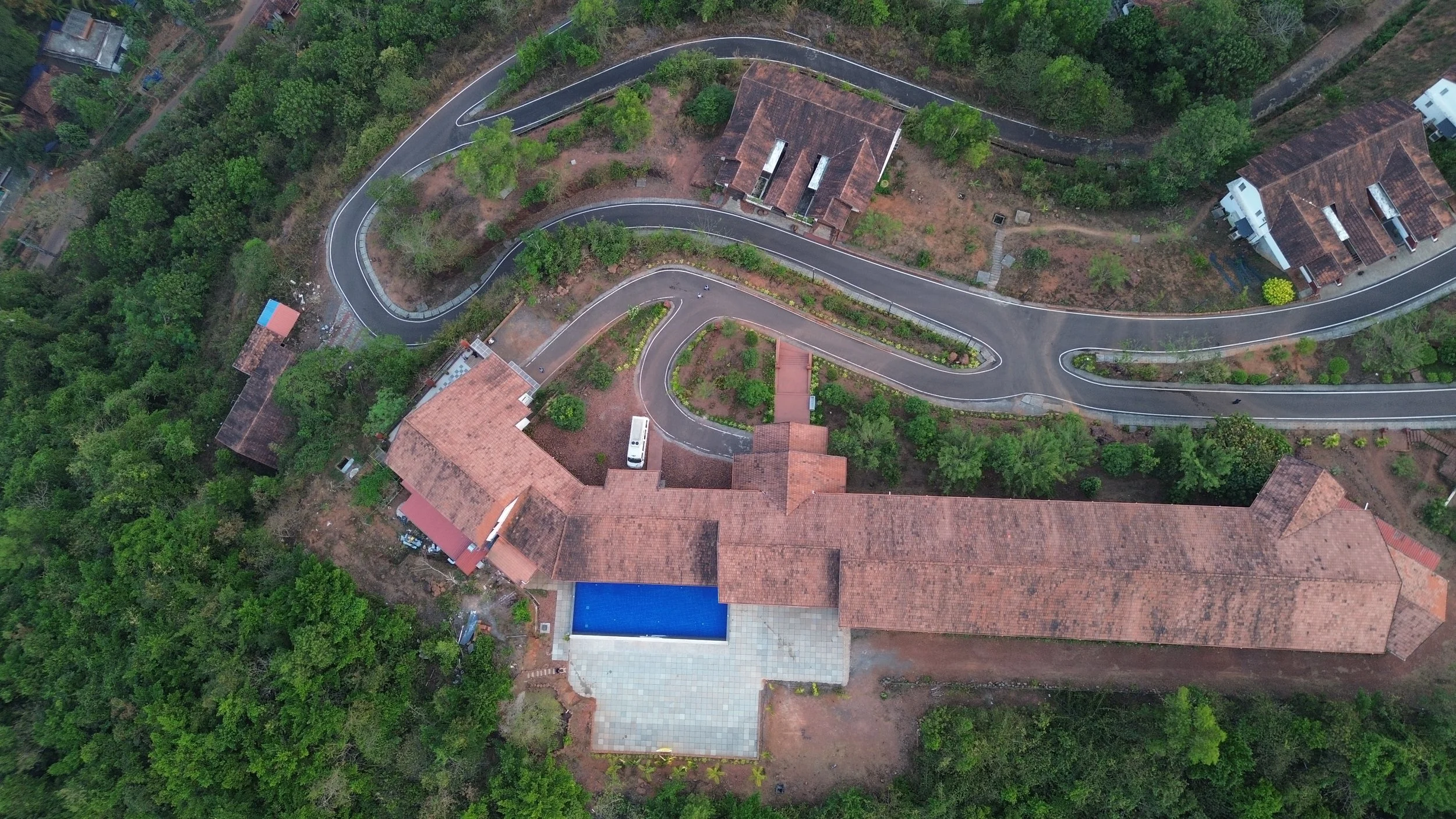 Aerial view of a large house with a swimming pool, surrounded by lush green trees and winding roads.