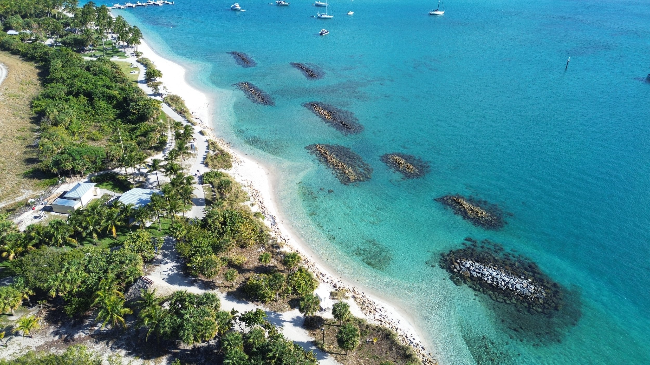 Aerial view of a tropical beach with white sand, palm trees, and turquoise water, featuring several rock formations and boats anchored offshore.