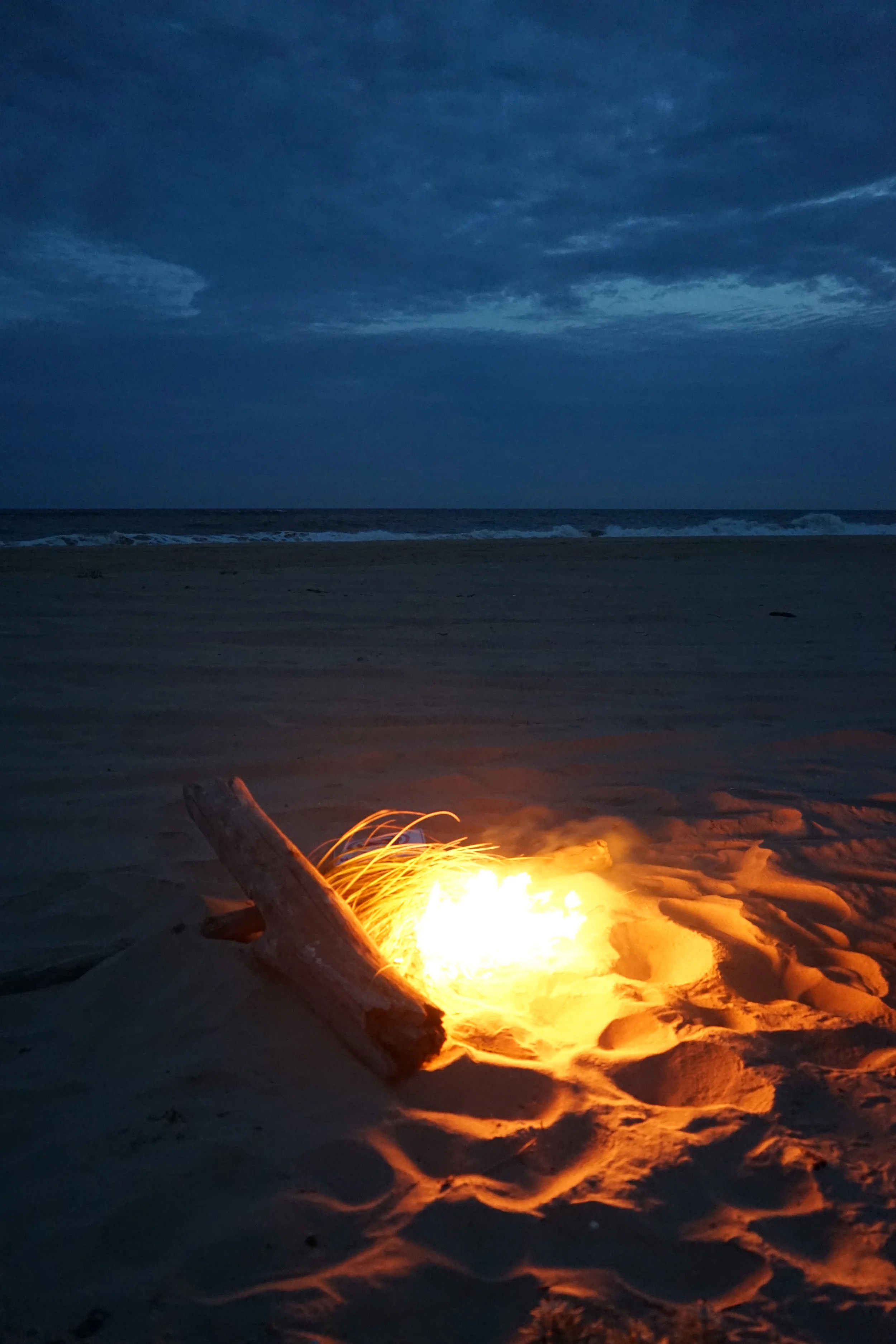 A campfire on a sandy beach at dusk, with the ocean and a cloudy sky in the background.