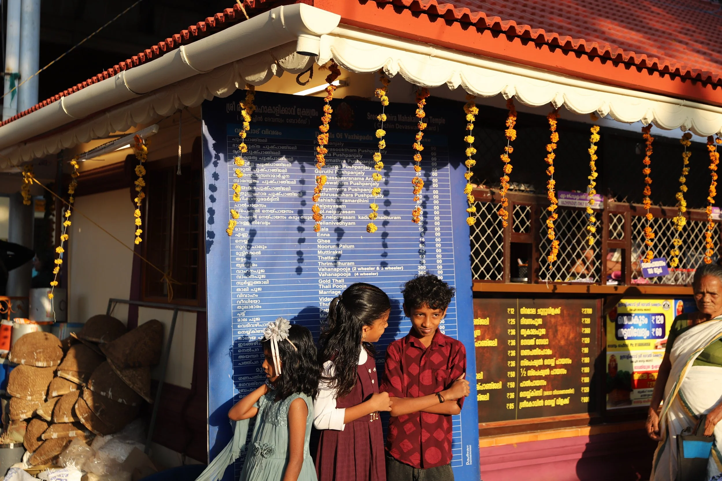 Three children standing in front of a blue menu board at an outdoor food stall decorated with marigold garlands, with a woman on the right side of the image.