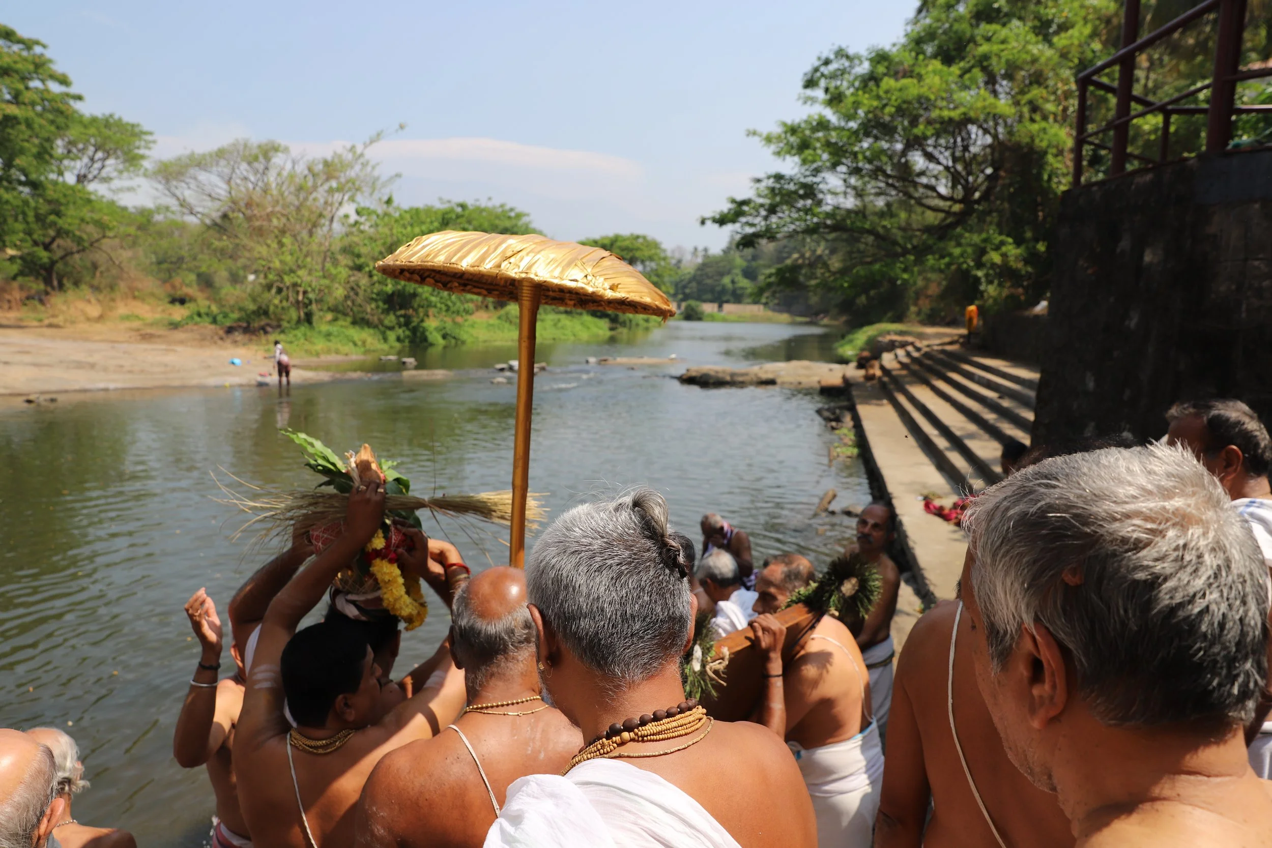 Hindu religious ceremony at a river with men dressed in traditional white dhotis, some with jewelry, performing rituals with offerings, flowers, and a parasol on a sunny day surrounded by trees and stone steps.