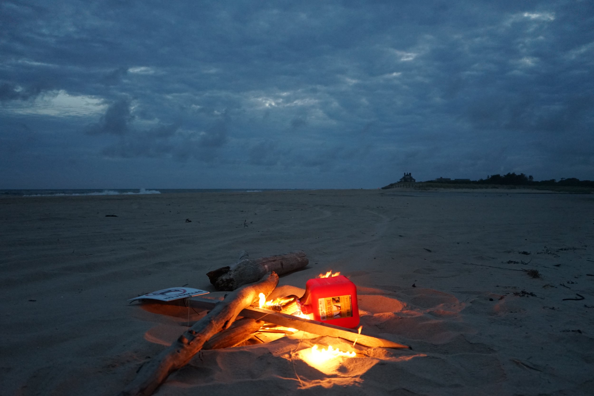 A small campfire burns on a sandy beach near driftwood and a red fuel container, with a cloudy sky overhead and a distant building on a hill in the background.