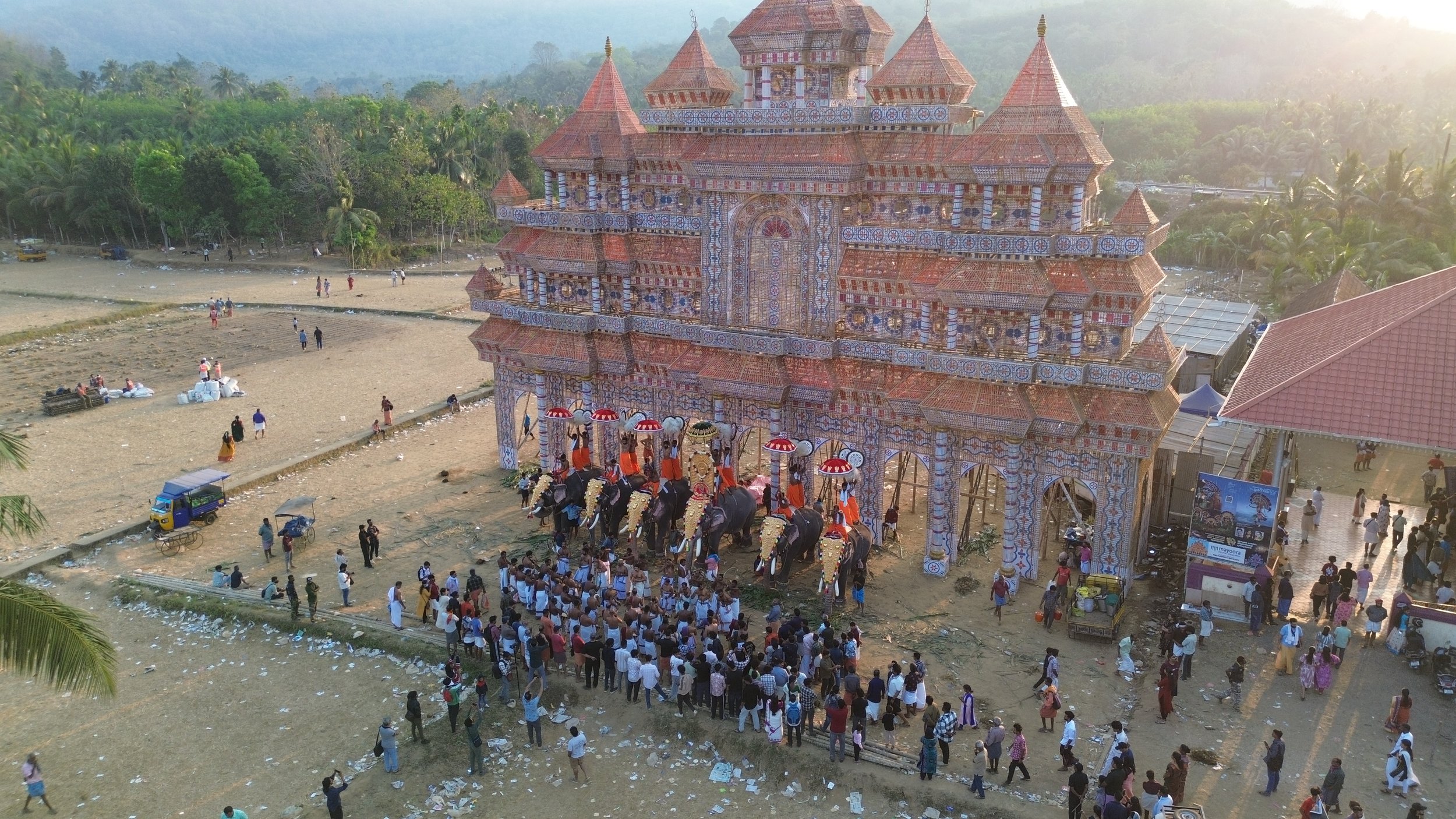 A large, ornate temple decorated with colorful patterns, with an elephant festival parade in front featuring elephants adorned with golden decorations and umbrellas, and a crowd of spectators watching.