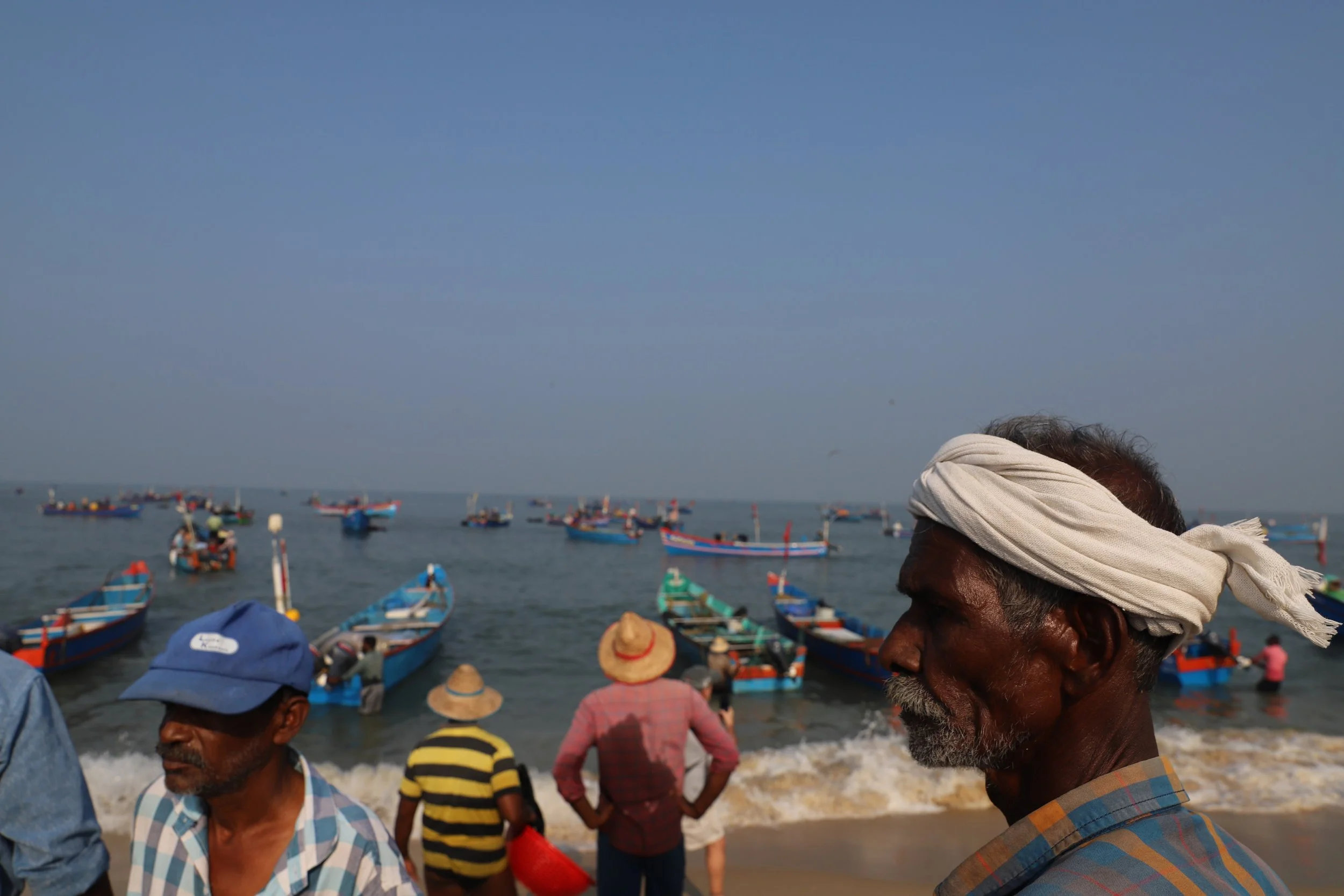 People on a beach with colorful boats in the water, some wearing straw hats and caps, under a clear blue sky.