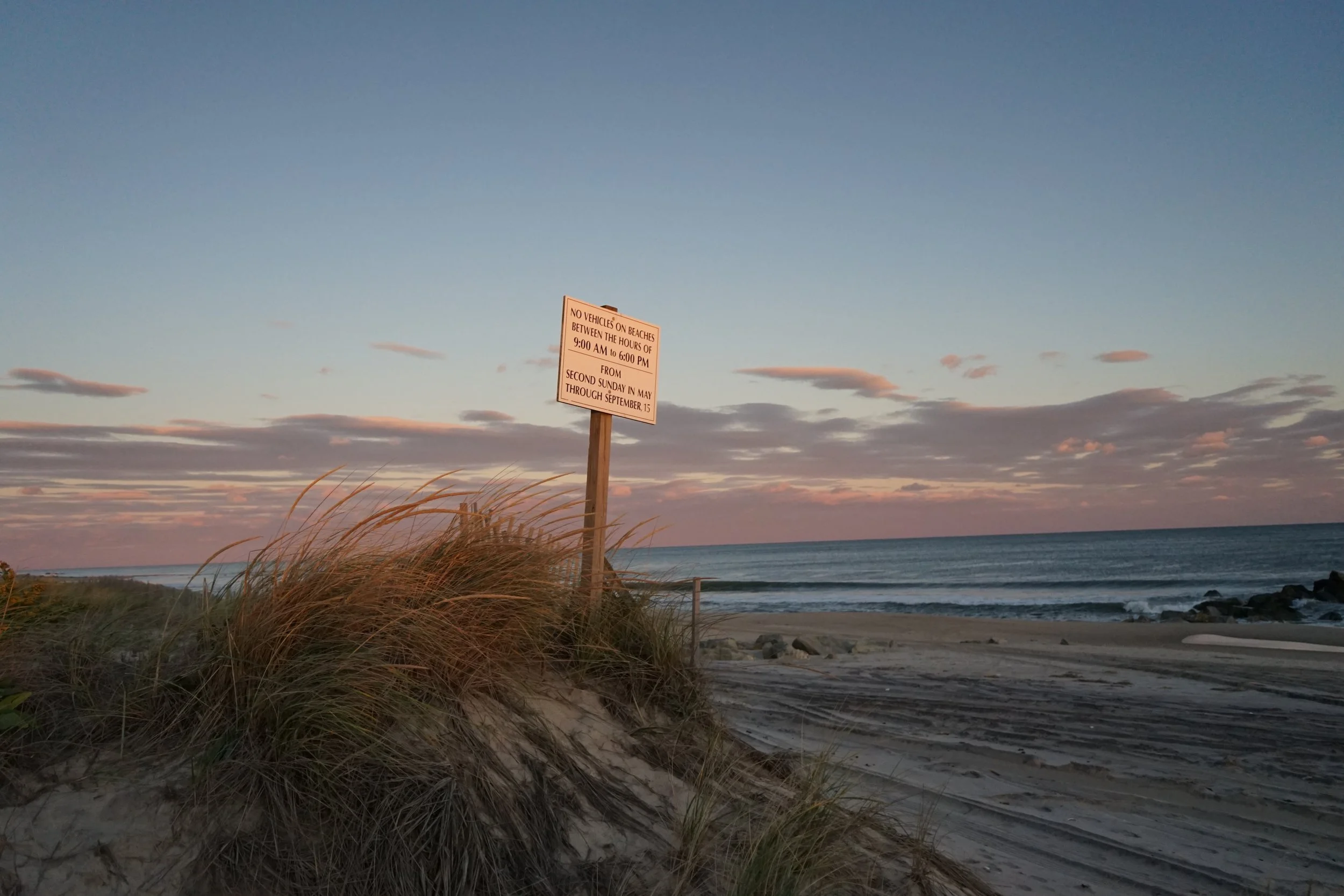 Beach with sand dunes and tall grasses, ocean waves, and a sunset sky with scattered clouds. A sign on a post indicates beach rules and seasonal regulations.