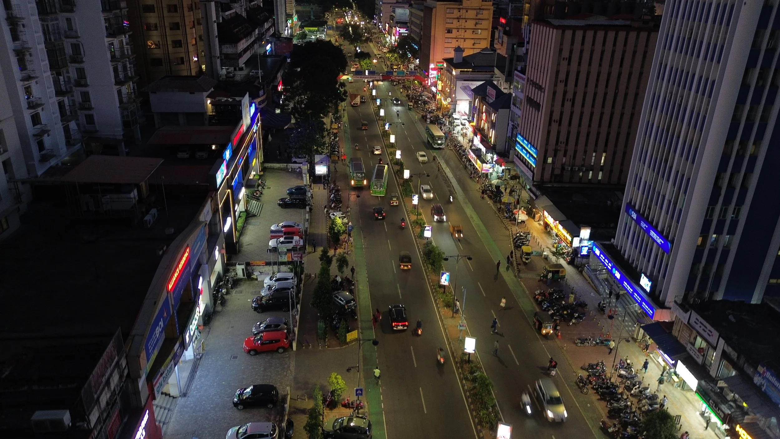 Nighttime city street shot from a high angle showing moving cars and motorcycles, parked vehicles, illuminated signs, buildings, and pedestrians.