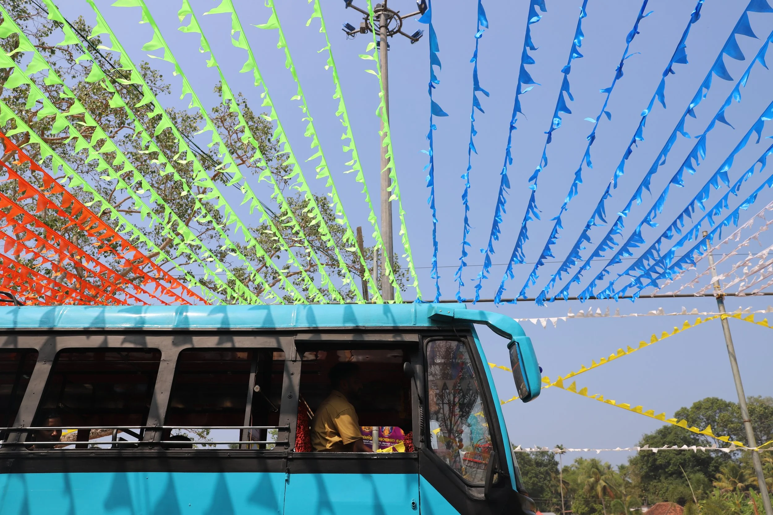 A blue vehicle with a person inside, parked under colorful decorative flags strung overhead during a festival or celebration on a clear day.