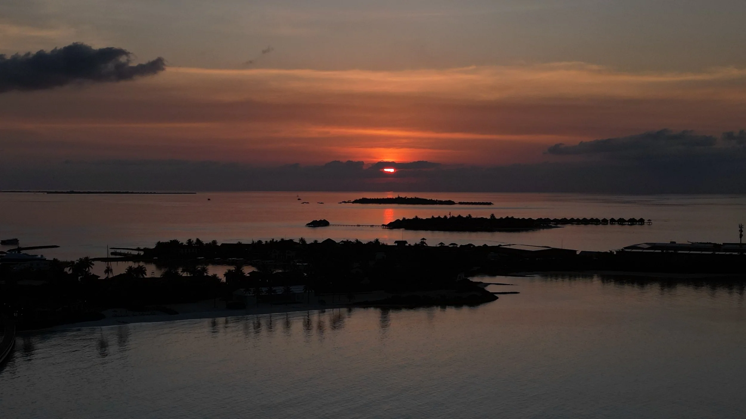 Sunset over a calm ocean with small islands and rocky outcroppings, and silhouettes of palm trees and buildings in the foreground.