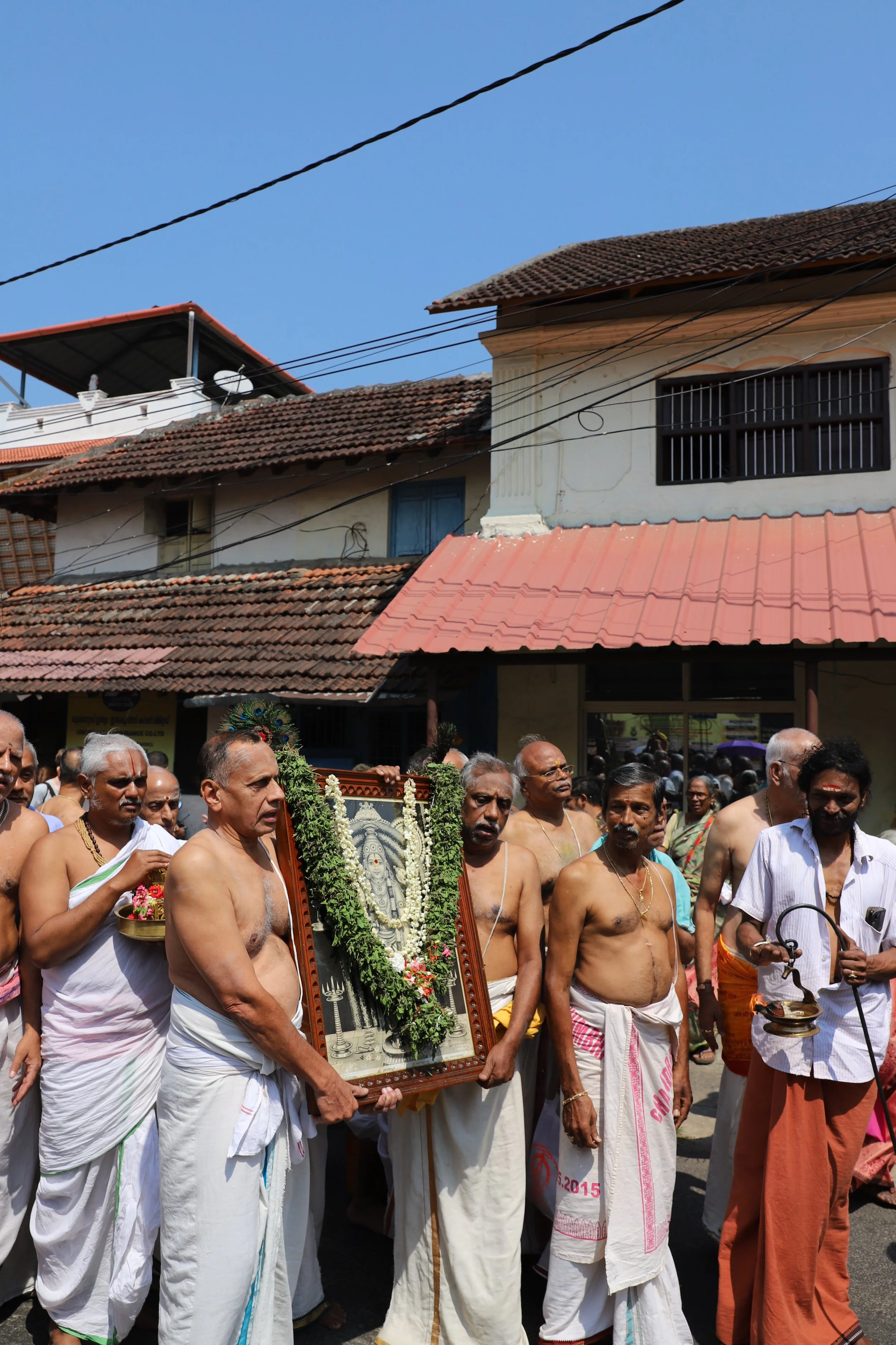 Hindu men participating in a religious procession, carrying a framed image of a deity decorated with flowers, surrounded by other men in traditional attire on a street in India.