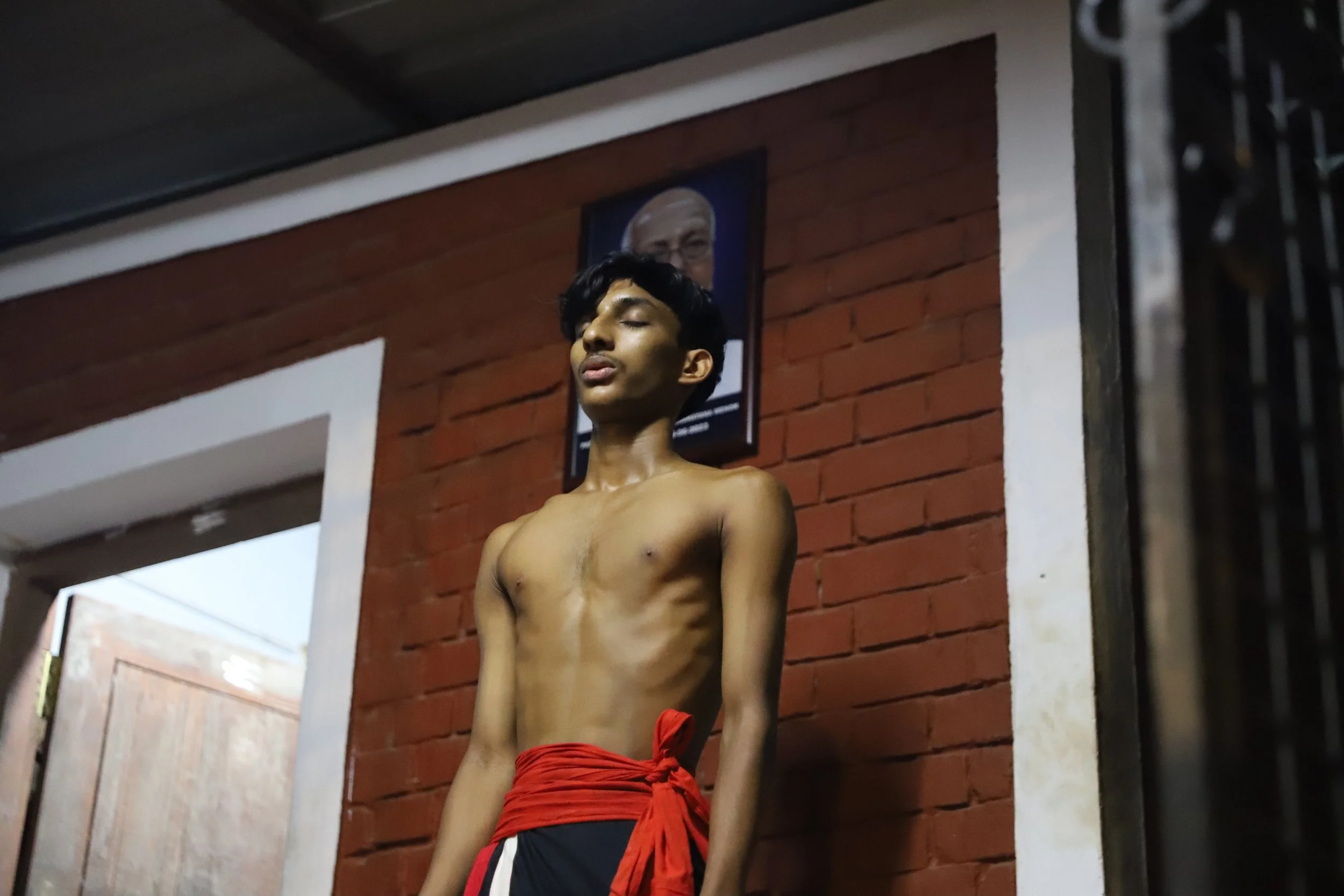 A young shirtless man with a red cloth tied around his waist stands with his eyes closed against a brick wall, with a framed photo of a man hanging above him.