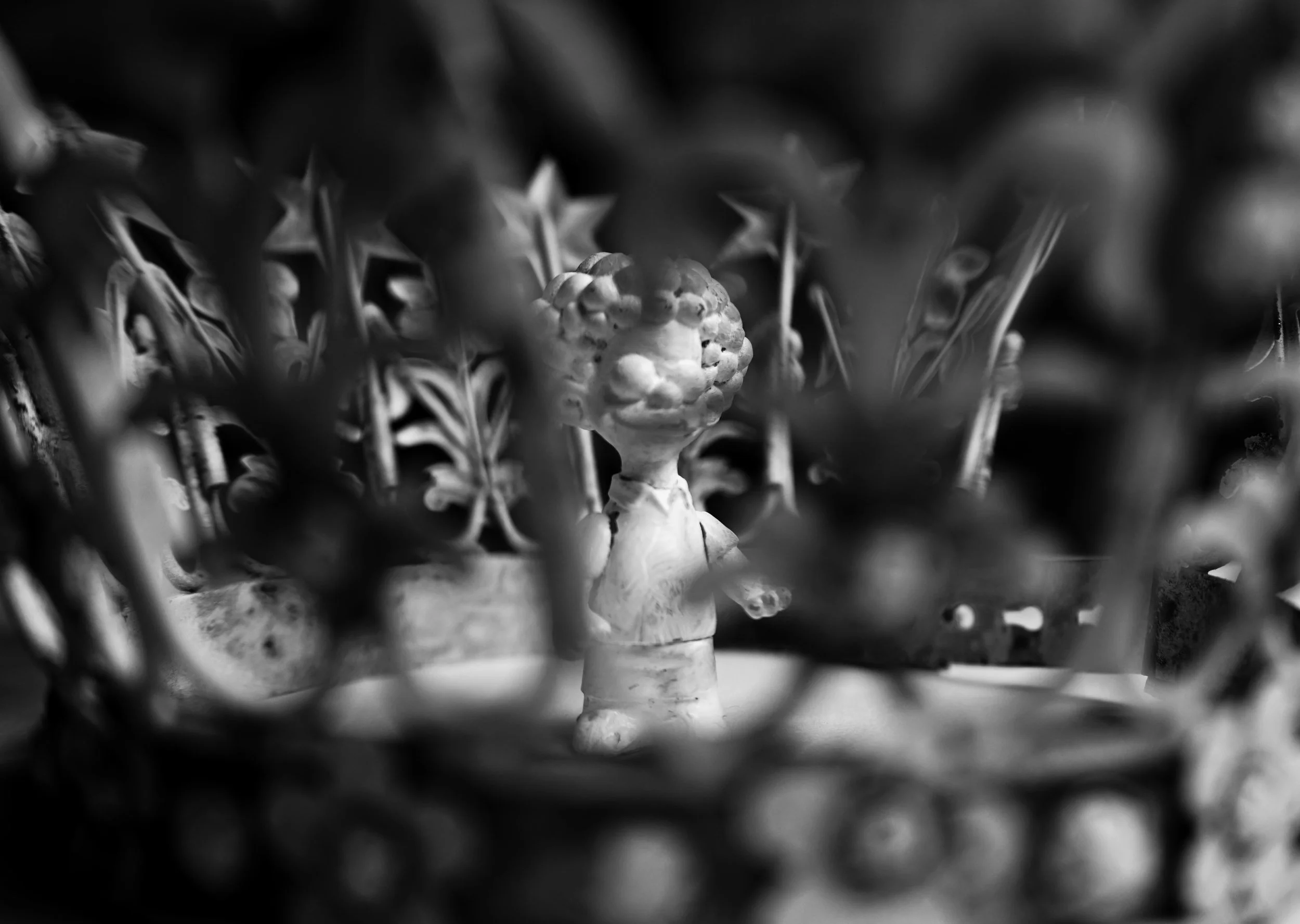 Black and white photo of a small figure with a head resembling a broccoli floret, standing among geometric patterned plants or decorations.