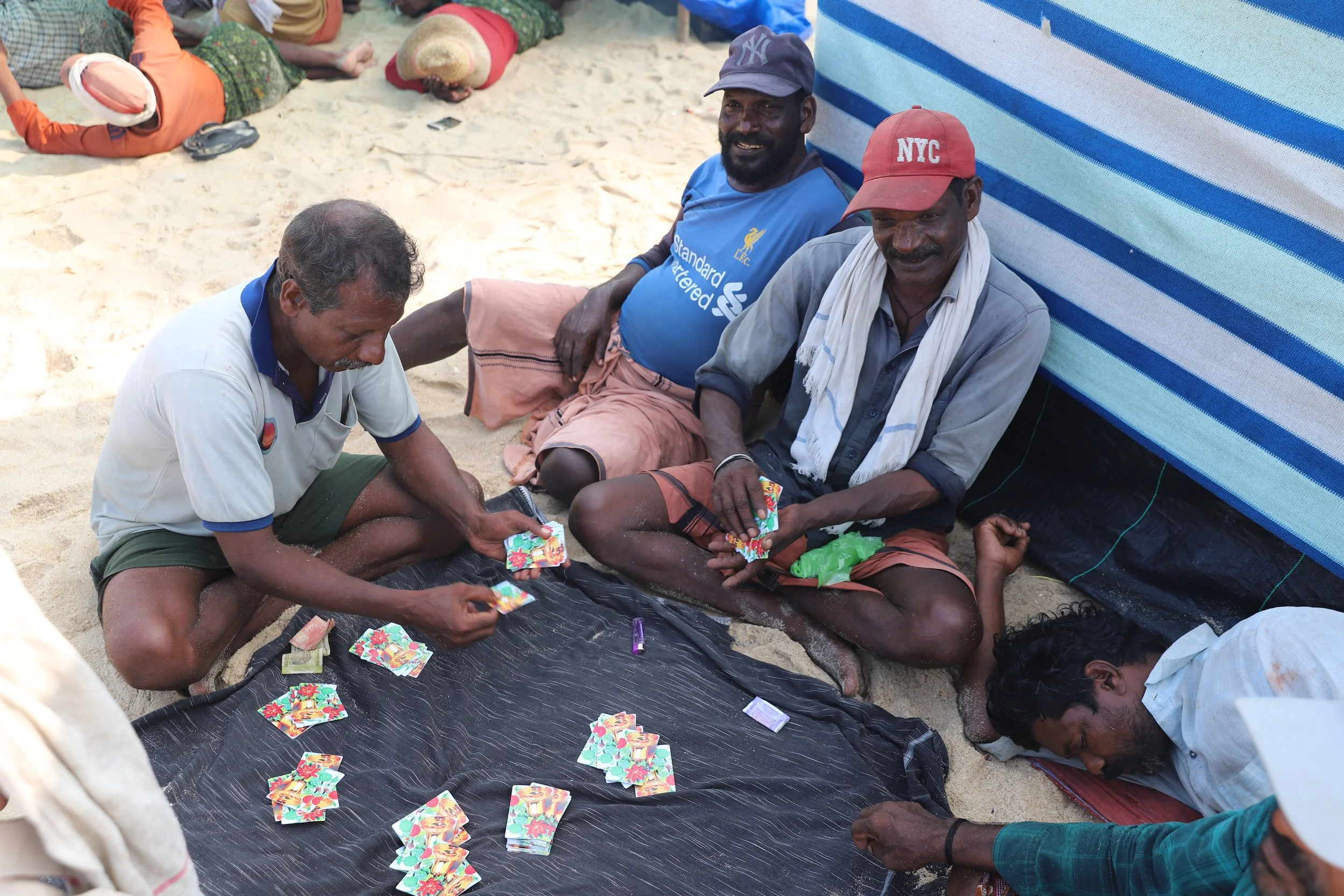 Group of men sitting on sand playing with colorful cards in front of a striped blue tent, with other people lying in the background.