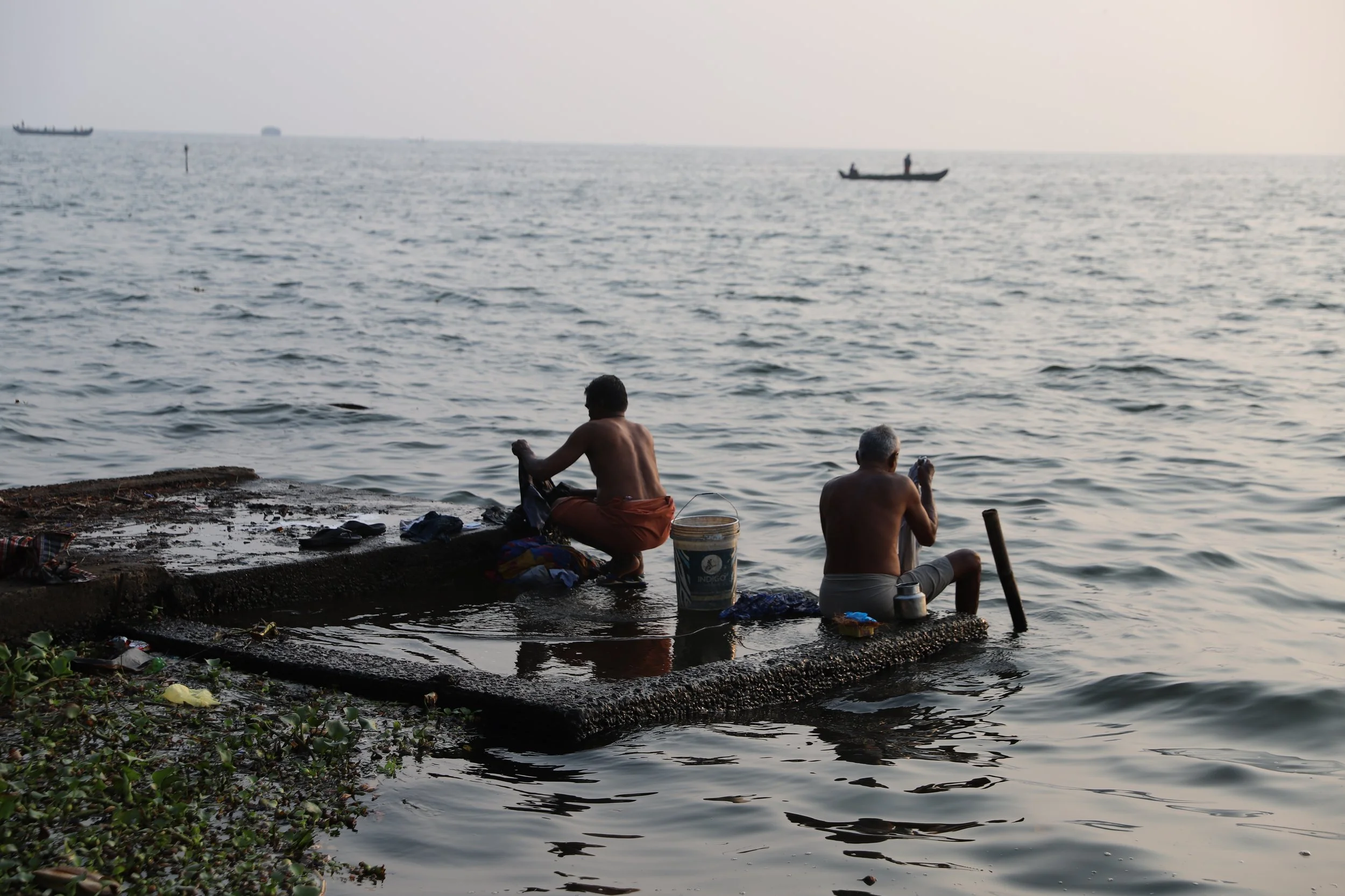 Two men sitting on a small concrete platform at the edge of a body of water, washing clothes with boats visible in the distance.