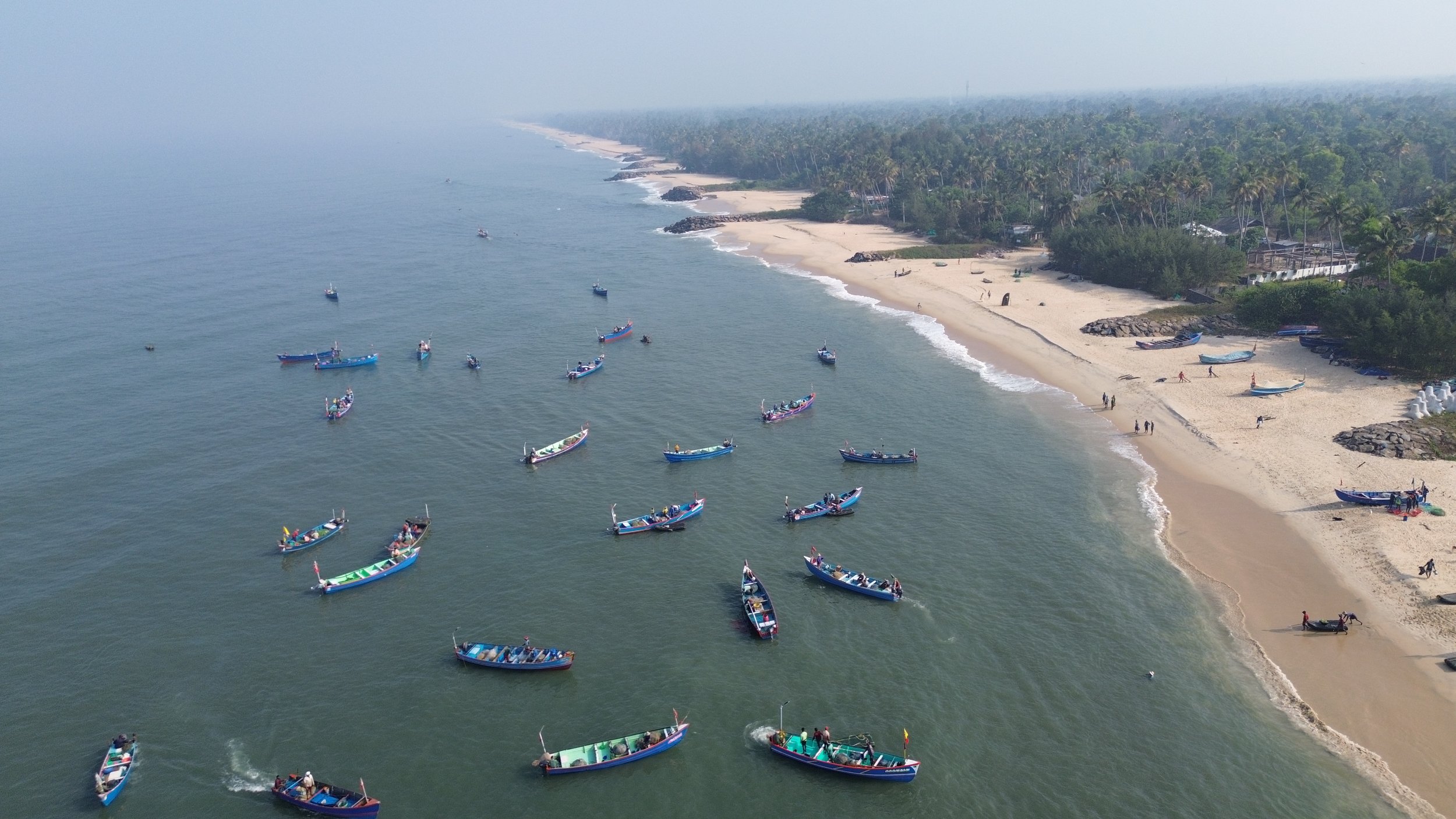 Aerial view of a coastal area with many small boats floating on the water near a sandy beach, with dense green vegetation and palm trees in the background.