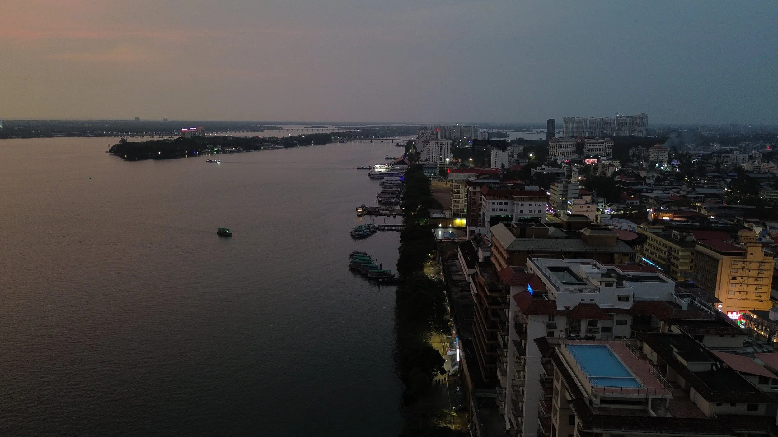 Cityscape view of a river at dusk with buildings and boats along the shoreline, some with lit pools and streetlights, under a cloudy sky.