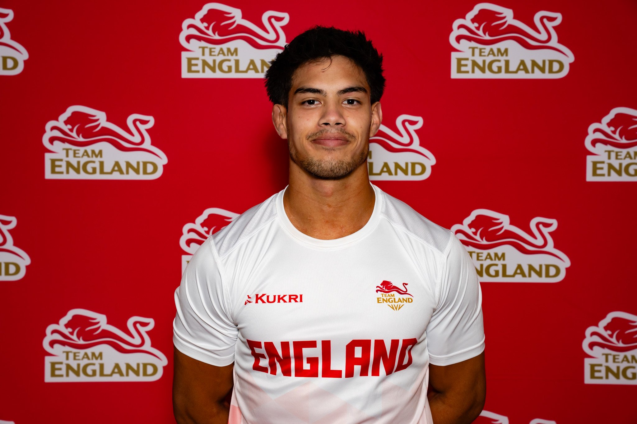 A young man standing in front of a red background with multiple 'Team England' logos. He is wearing a white sports shirt with 'Kukri' and 'England' logos.