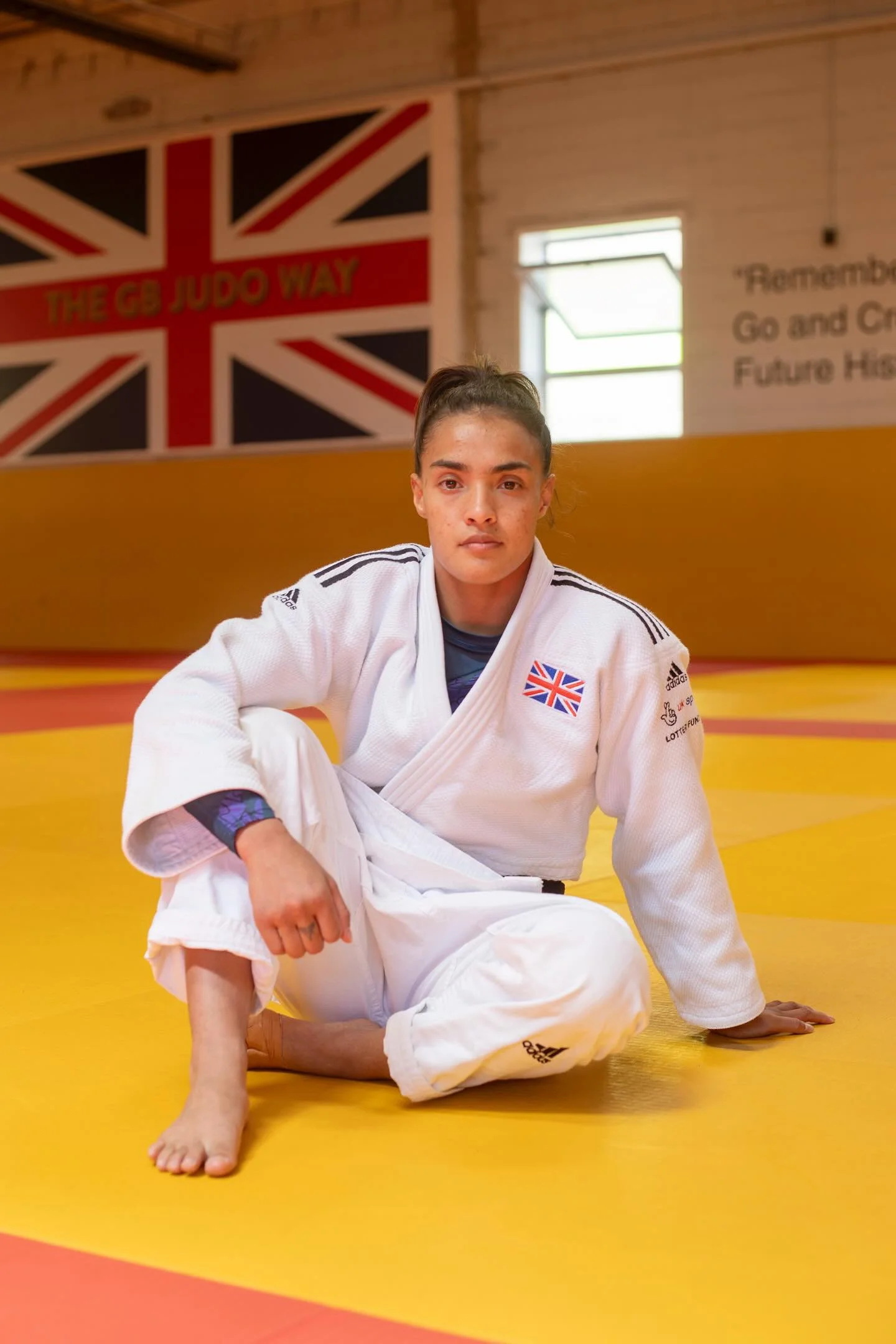 A woman in a white judo gi with a British flag patch on her chest sitting on her knees with one hand resting on the mat in a dojo, with a British flag and motivational text on the wall behind her.