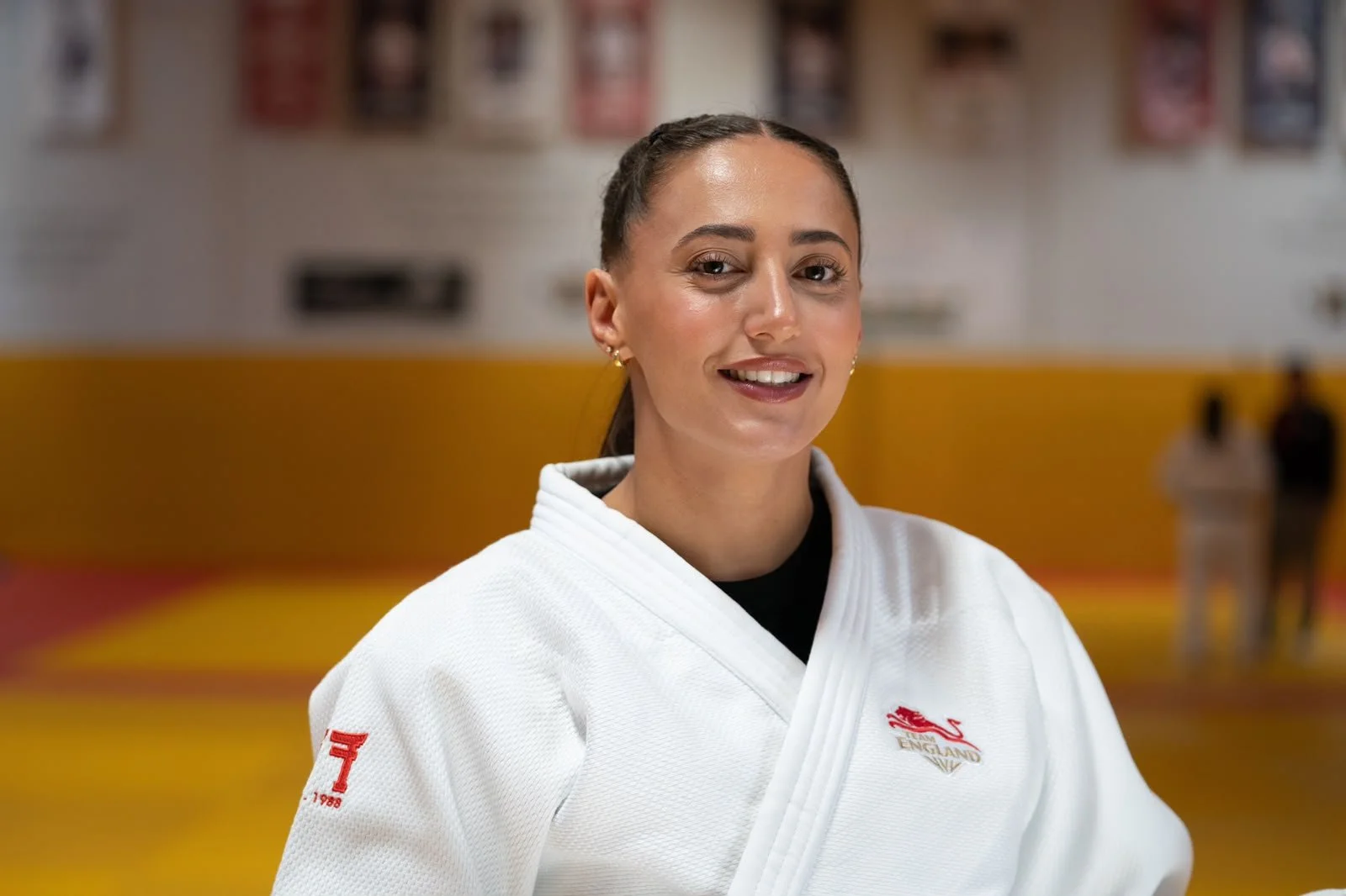 Shelley Ludford in a white judo gi with England team badges stands on a yellow and red judo mat, with banners and people in the background.