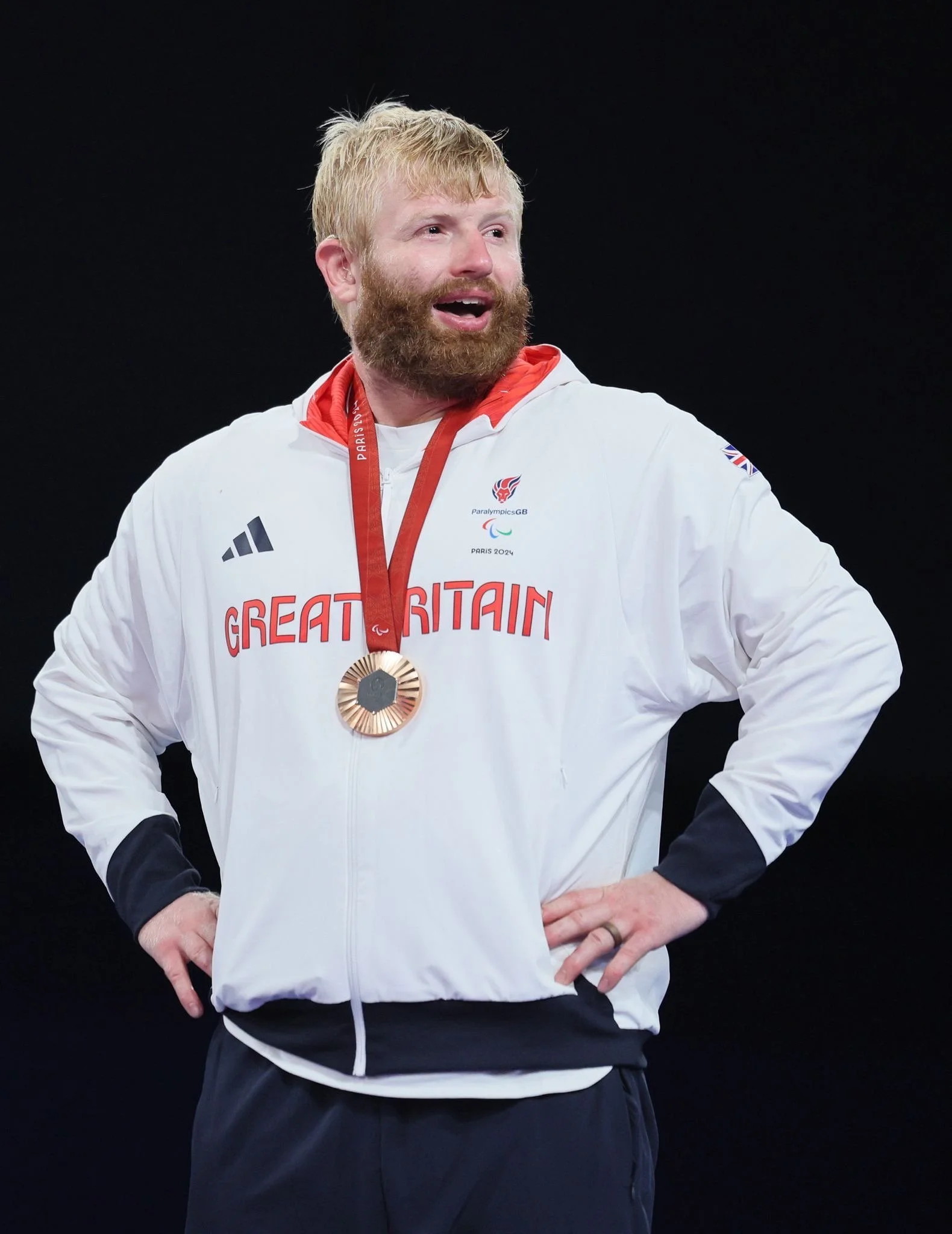 A man with red hair and a beard wearing a white jacket with a medal around his neck, standing against a black background.