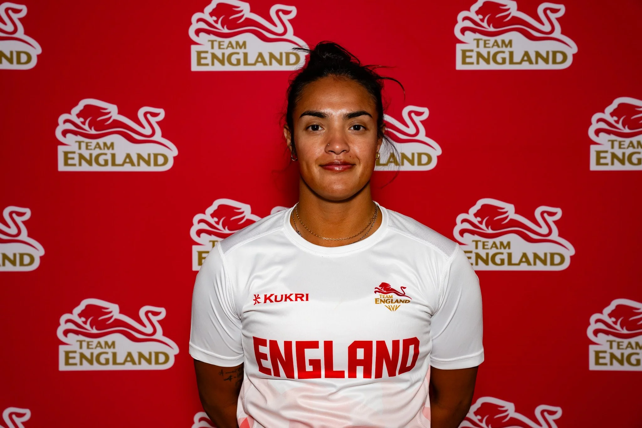 Lele NAIRNE standing in front of a red background with 'Team England' logos, wearing a white sports jersey with 'England' written on it and a small 'Kukri' logo.
