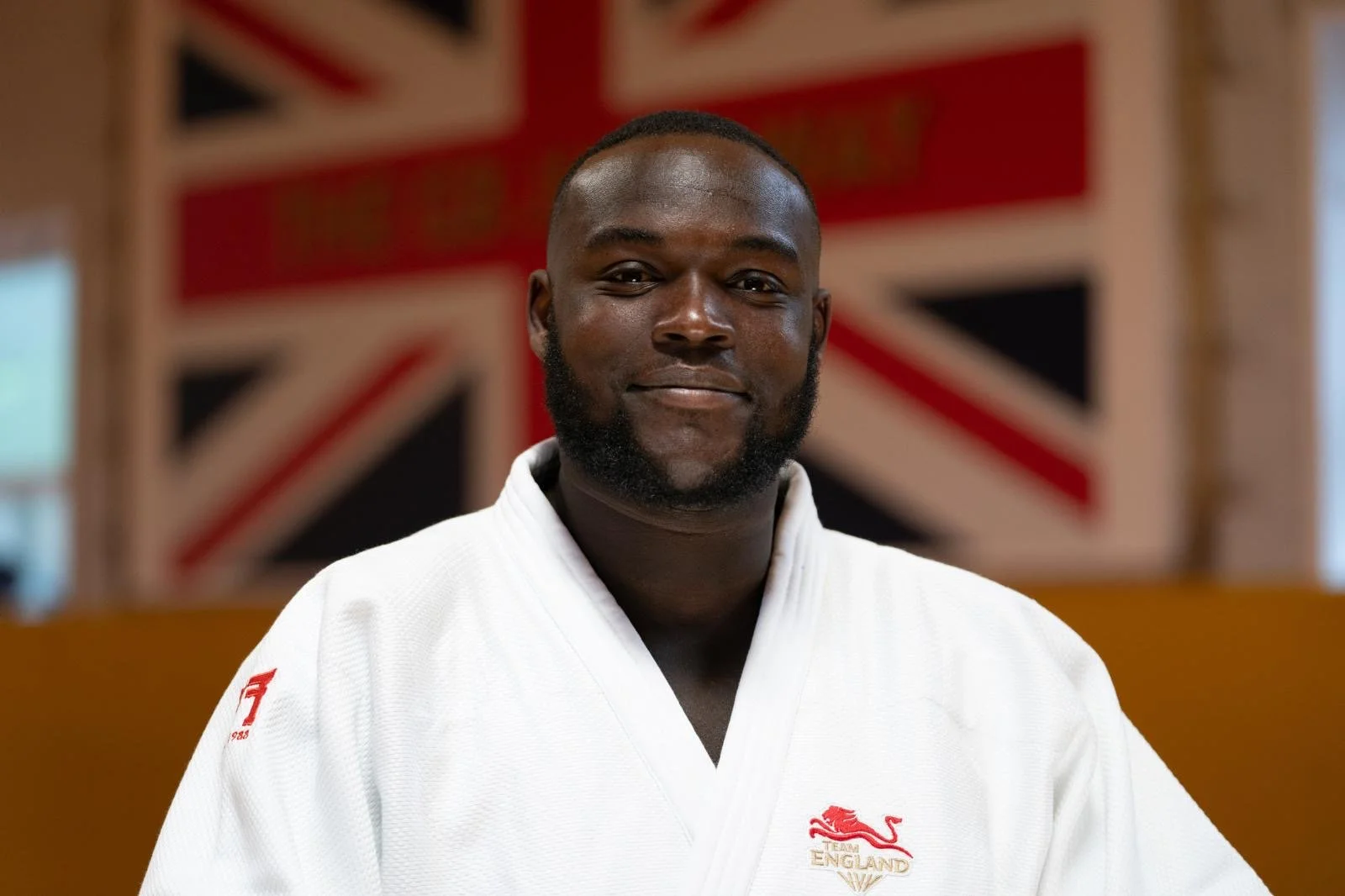 A man with a beard and short hair wearing a white judo gi with an England team logo, standing in front of a British flag background.