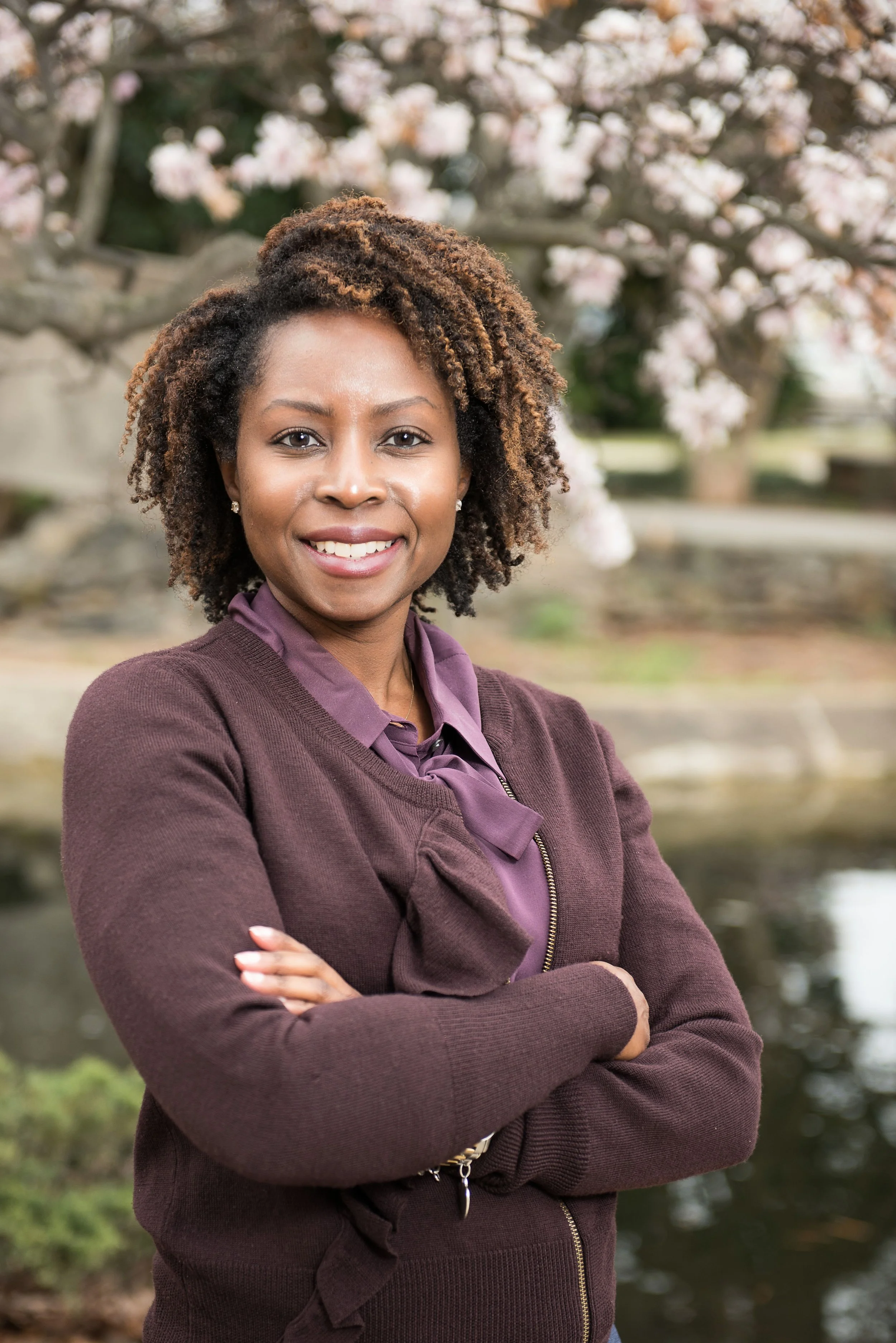 A Black woman with natural curly hair, wearing a purple blouse and a maroon sweater, standing outdoors with arms crossed. Cherry blossoms and a pond are in the background.