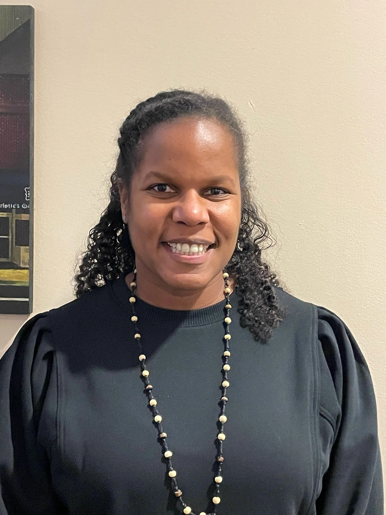 A Black woman with curly hair wearing a black top with puffy sleeves and a beaded necklace, smiling indoors against a plain wall.