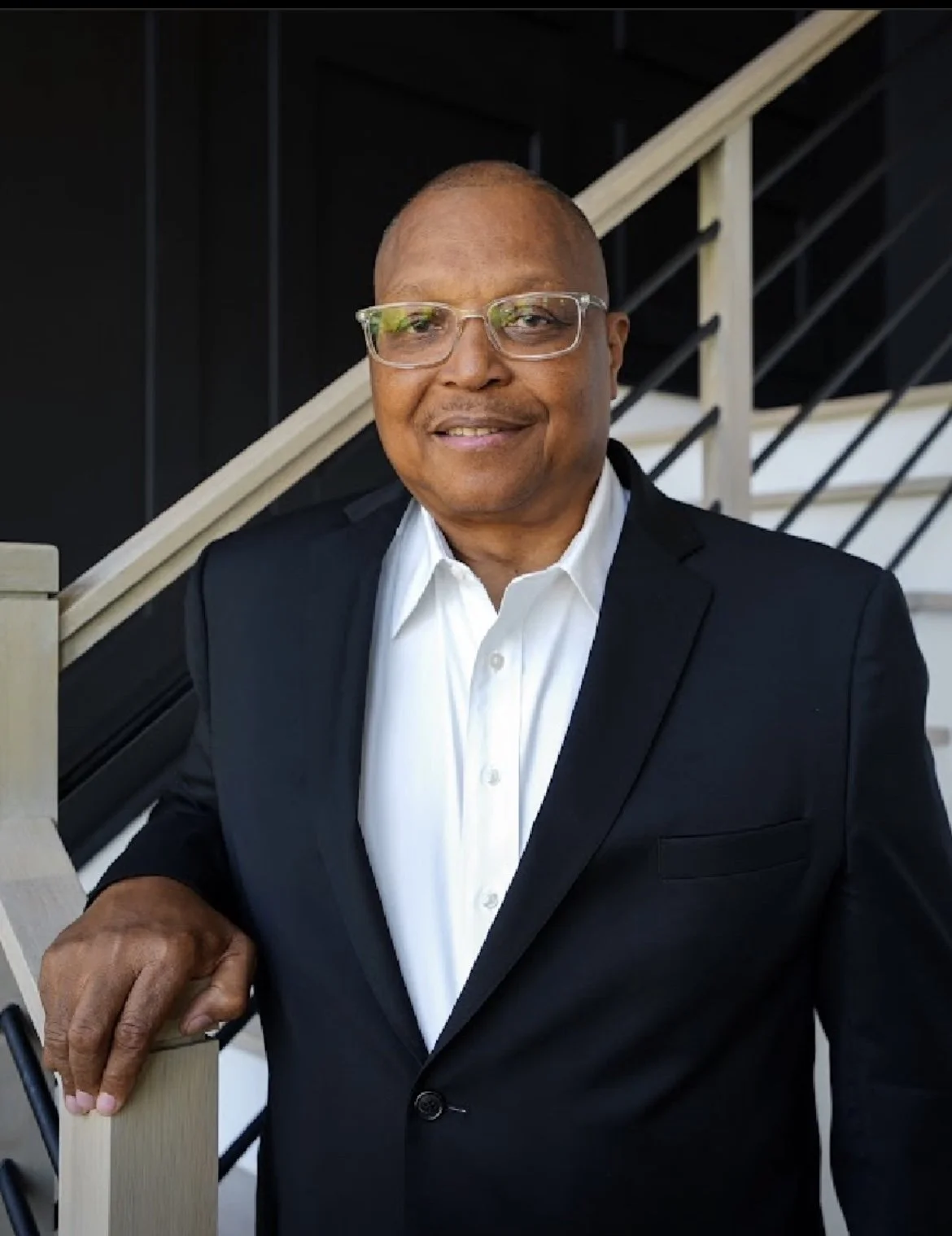 A Black man in a black suit and white shirt standing on a staircase with a railing.