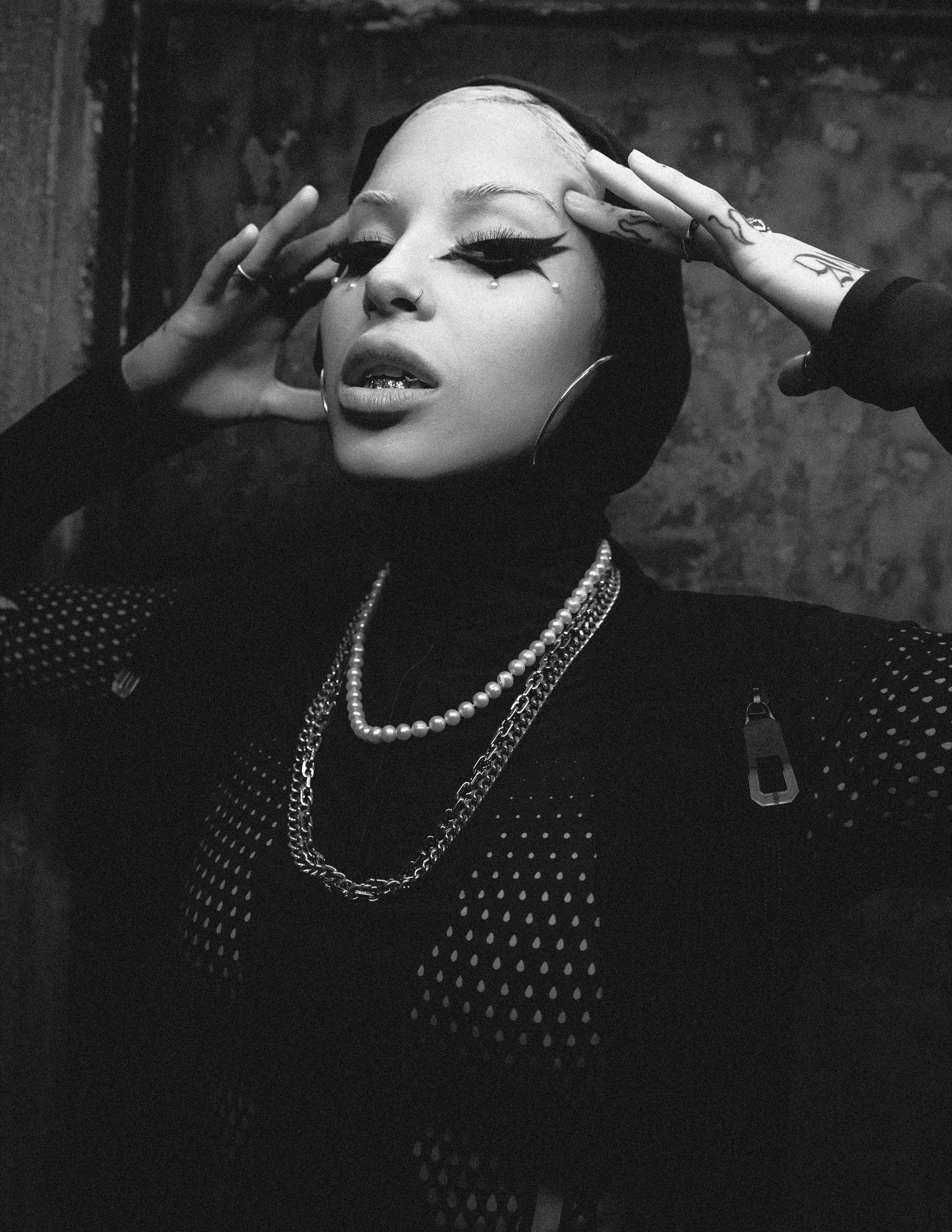 A woman with eye makeup, piercings, tattoos, and jewelry poses with her hands framing her face against a textured dark background.