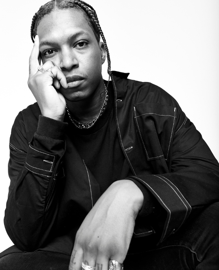 Black and white portrait of a person with braided hair, wearing a black long-sleeve shirt with visible stitching, posing with one hand resting on their face, gazing directly at the camera.