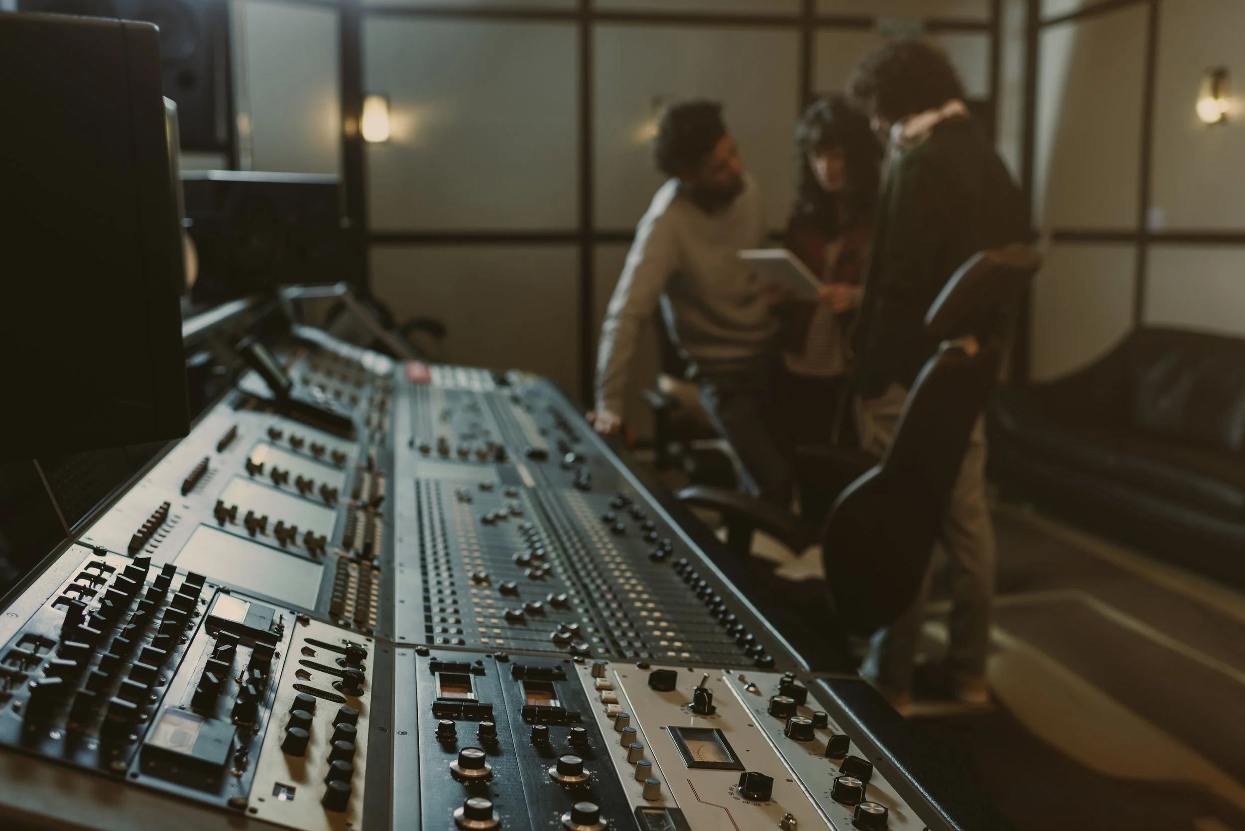 Recording studio with sound mixing console and three people reviewing notes in the background.