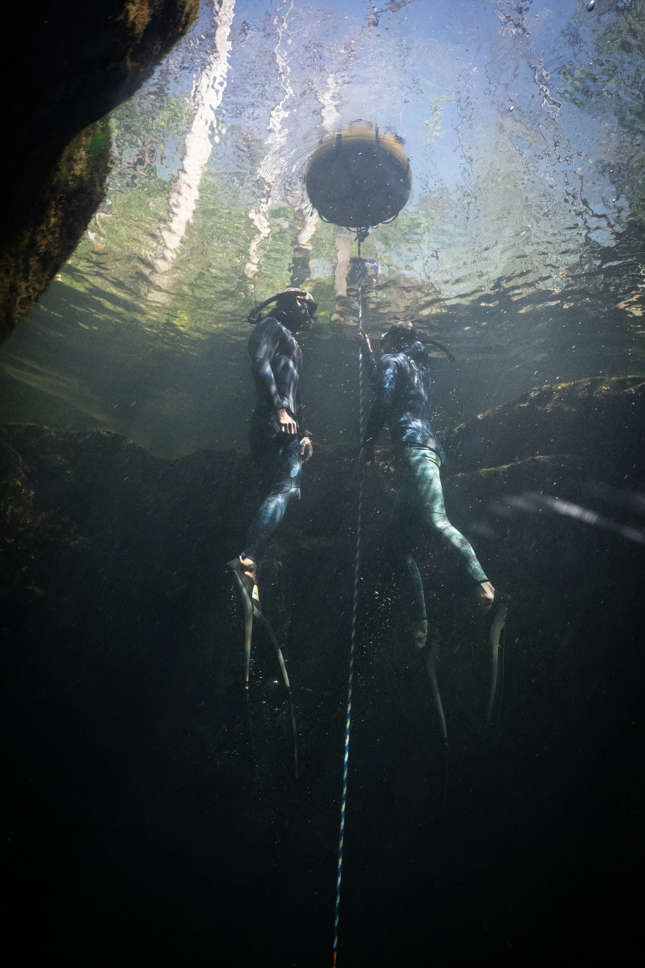 Two freedivers underwater near a cavern ceiling, holding a navigation line, with one diver reaching toward a float attached above them.