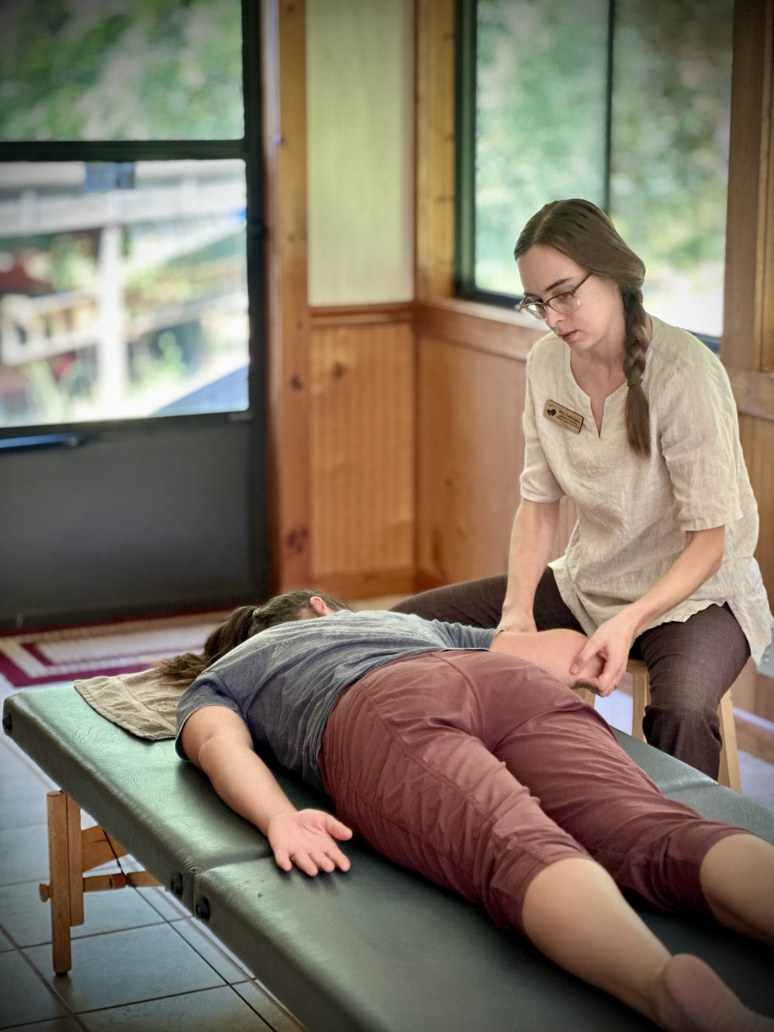 A woman receiving a massage from a massage therapist in a cozy wooden room with large windows showing a green outdoor scene.