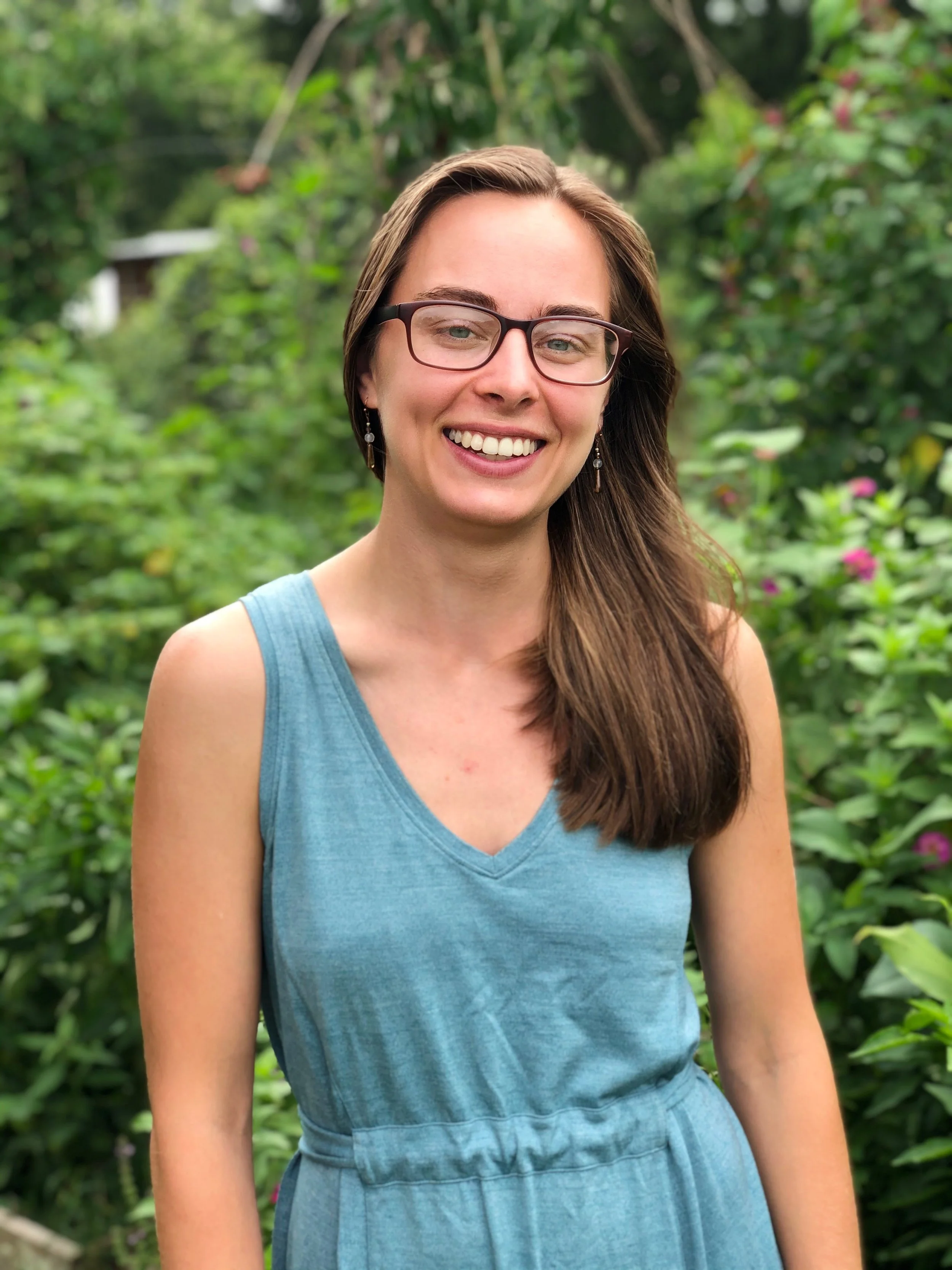 Smiling woman with brown hair and glasses standing outdoors in a lush green garden.