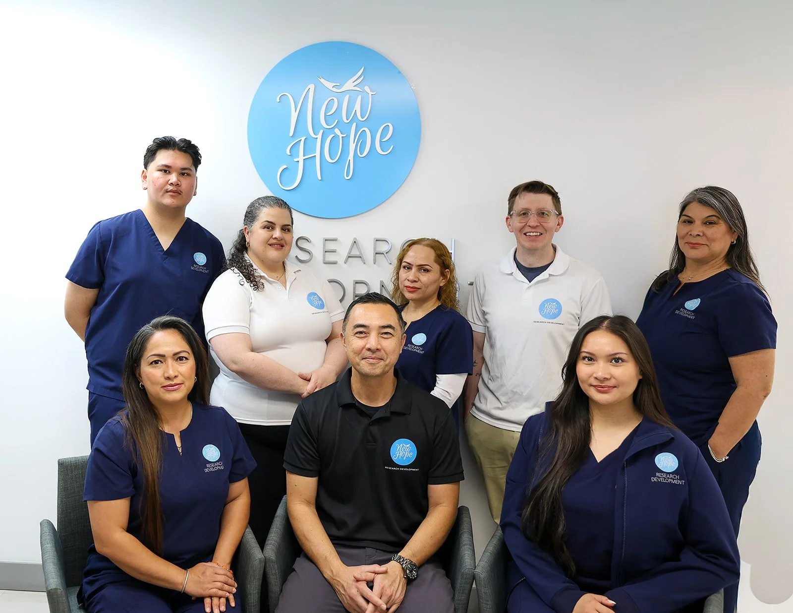 Group of nine diverse individuals, some in medical scrubs and some in casual white t-shirts with 'Research and Development' logo, posing for a photo in front of a wall with a 'New Hope' logo, in a professional setting.