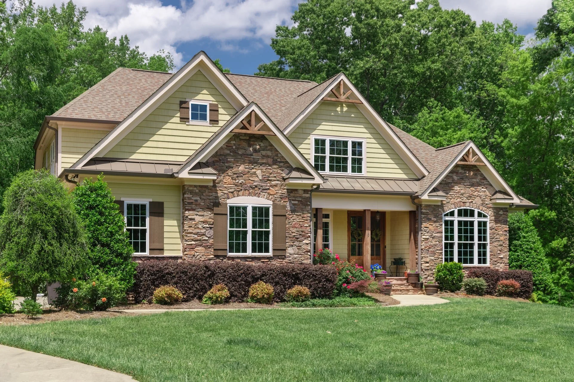 A large house with a mix of yellow siding and stone exterior, multiple gable roofs, and white framed windows. The house is surrounded by well-maintained green grass and shrubs, with tall trees in the background under a partly cloudy sky.