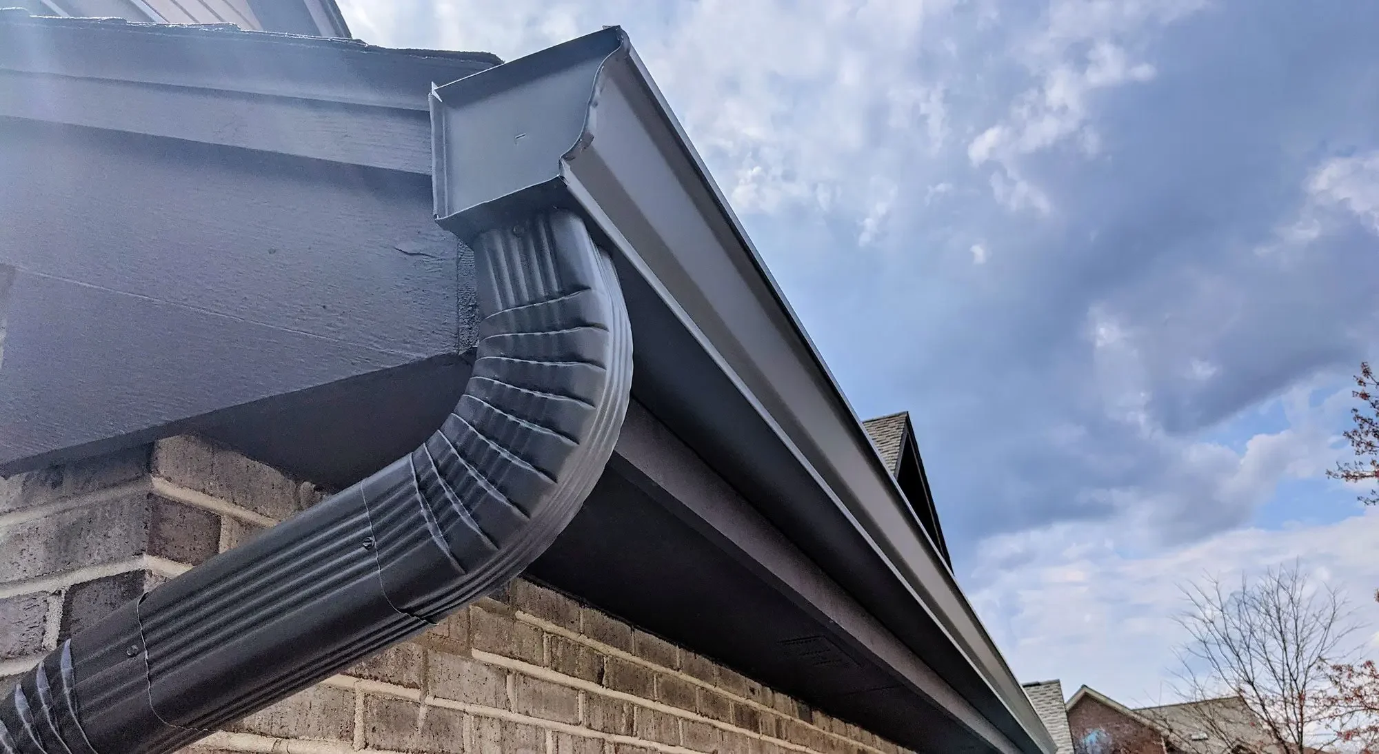 Close-up of a house's brick exterior, showing a black gutter connected to a downspout. The gutter and downspout are partially covered with a flexible, ribbed black protective sleeve. The sky is partly cloudy, with rooftops of neighboring houses visible in the background.