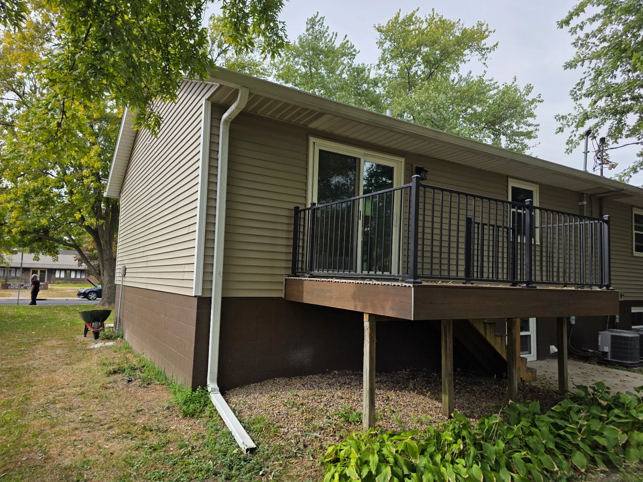 Back of a beige house with a small elevated wooden deck and black metal railing, surrounded by green trees and grass.