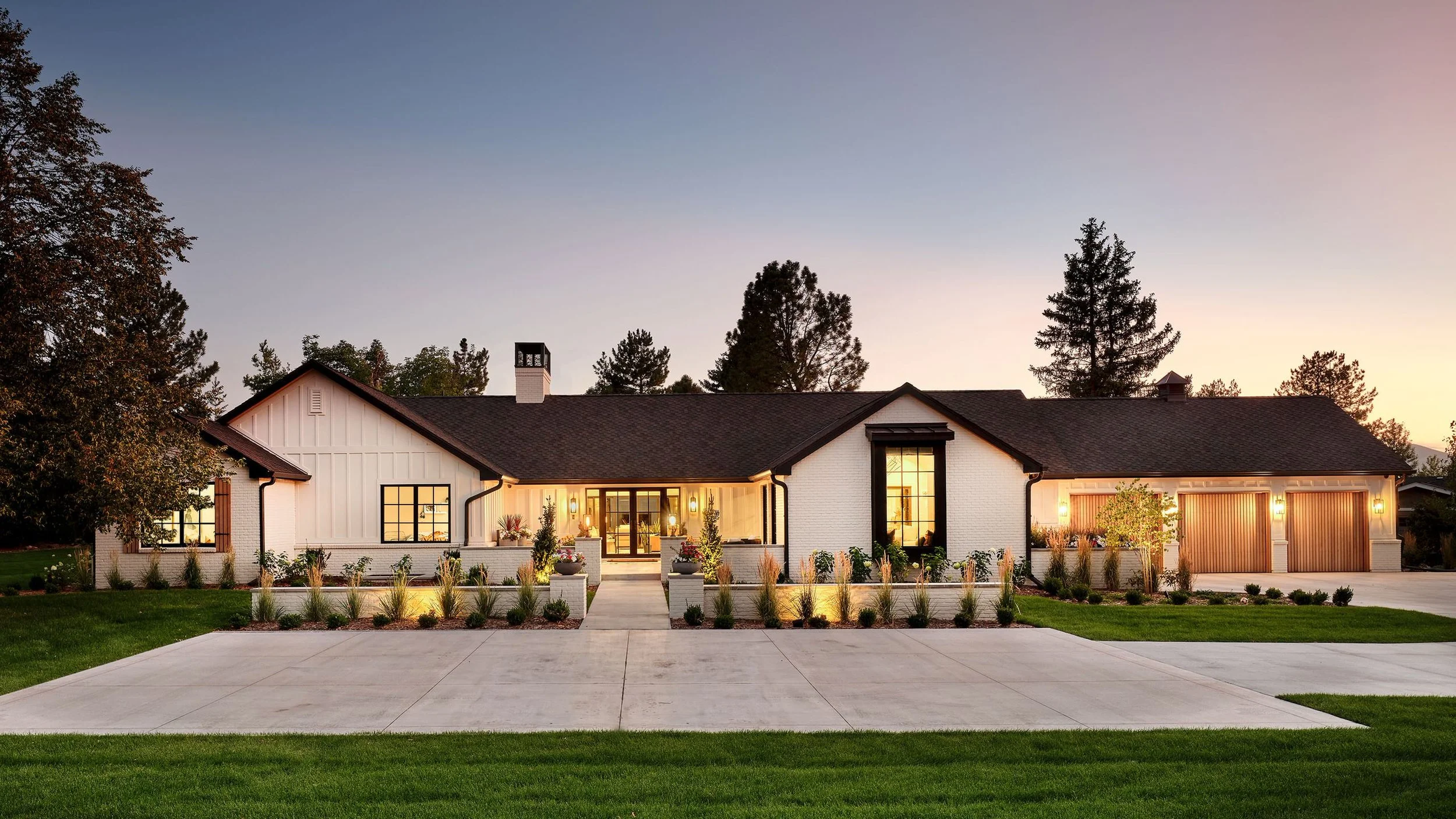 Front view of a modern suburban house during sunset, with a well-maintained lawn, flower beds, and driveway.