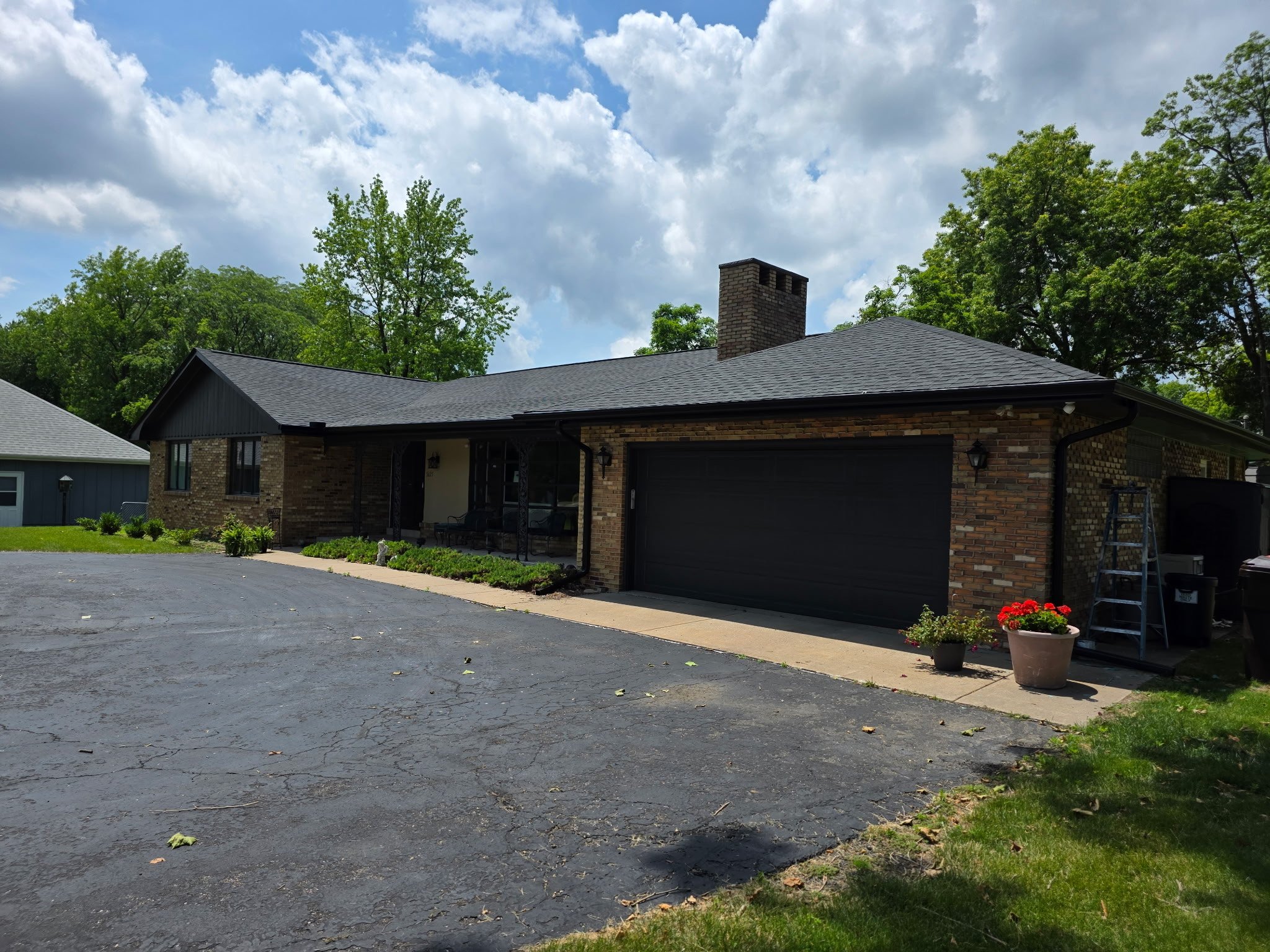 Single-story house with brick exterior, black garage door, and chimney, surrounded by trees and a landscaped front yard.
