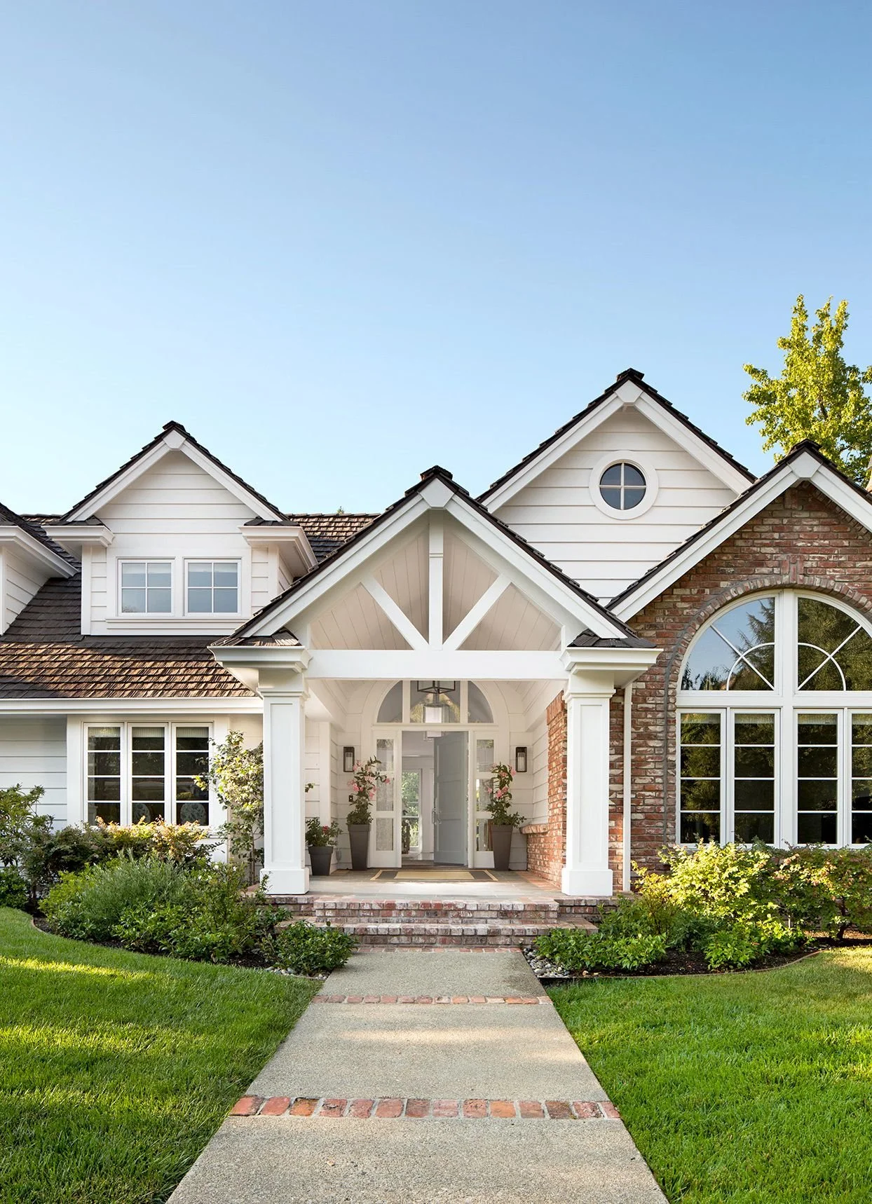Front view of a modern house with white siding, brick accents, and large arched windows surrounded by green landscaping and a paved walkway leading to the front door.