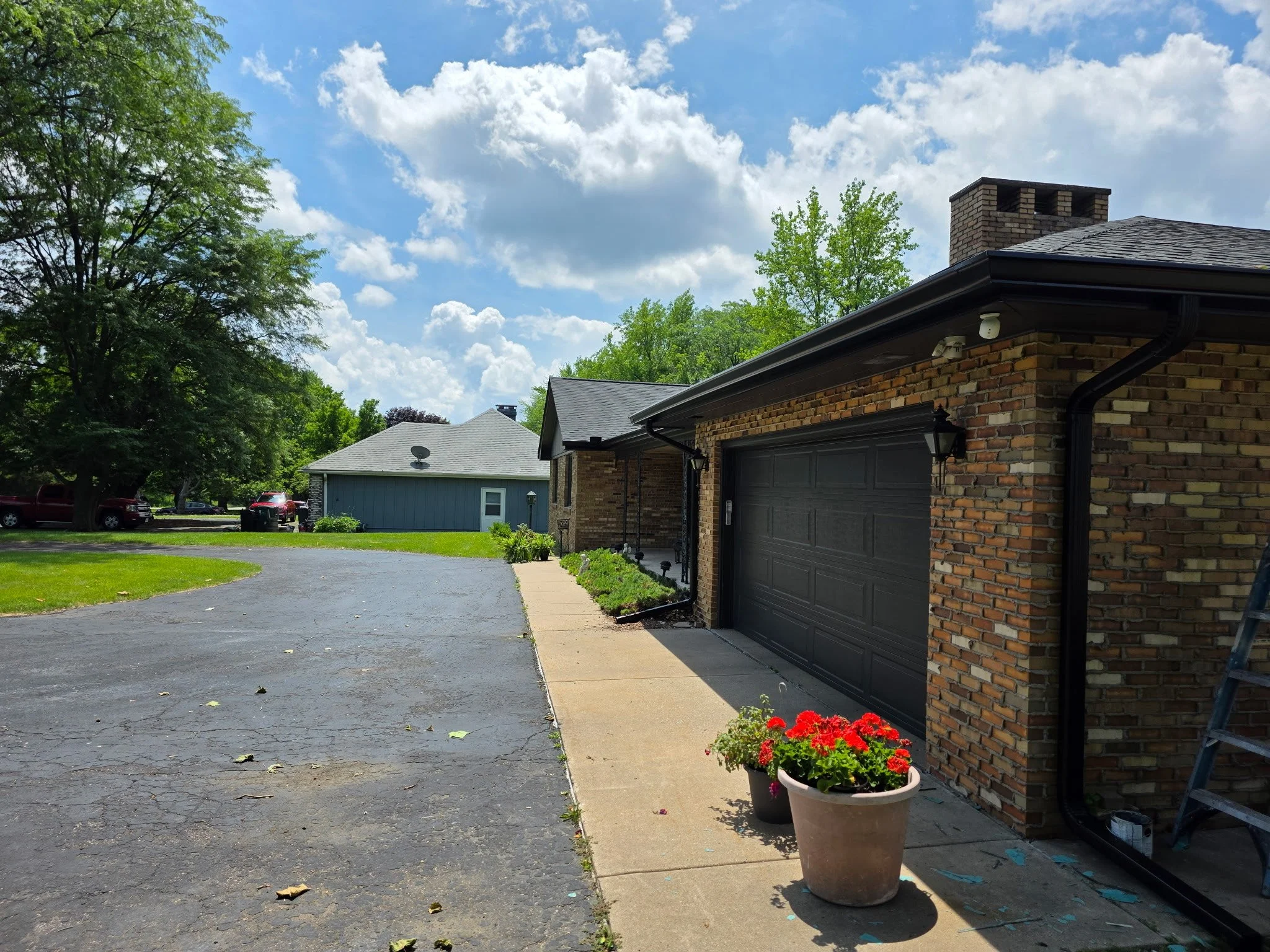 A brick house with a closed garage door, potted red flowers, and a sidewalk in front. A paved driveway curves to the left, leading to other houses in a suburban neighborhood with green trees and blue sky overhead.