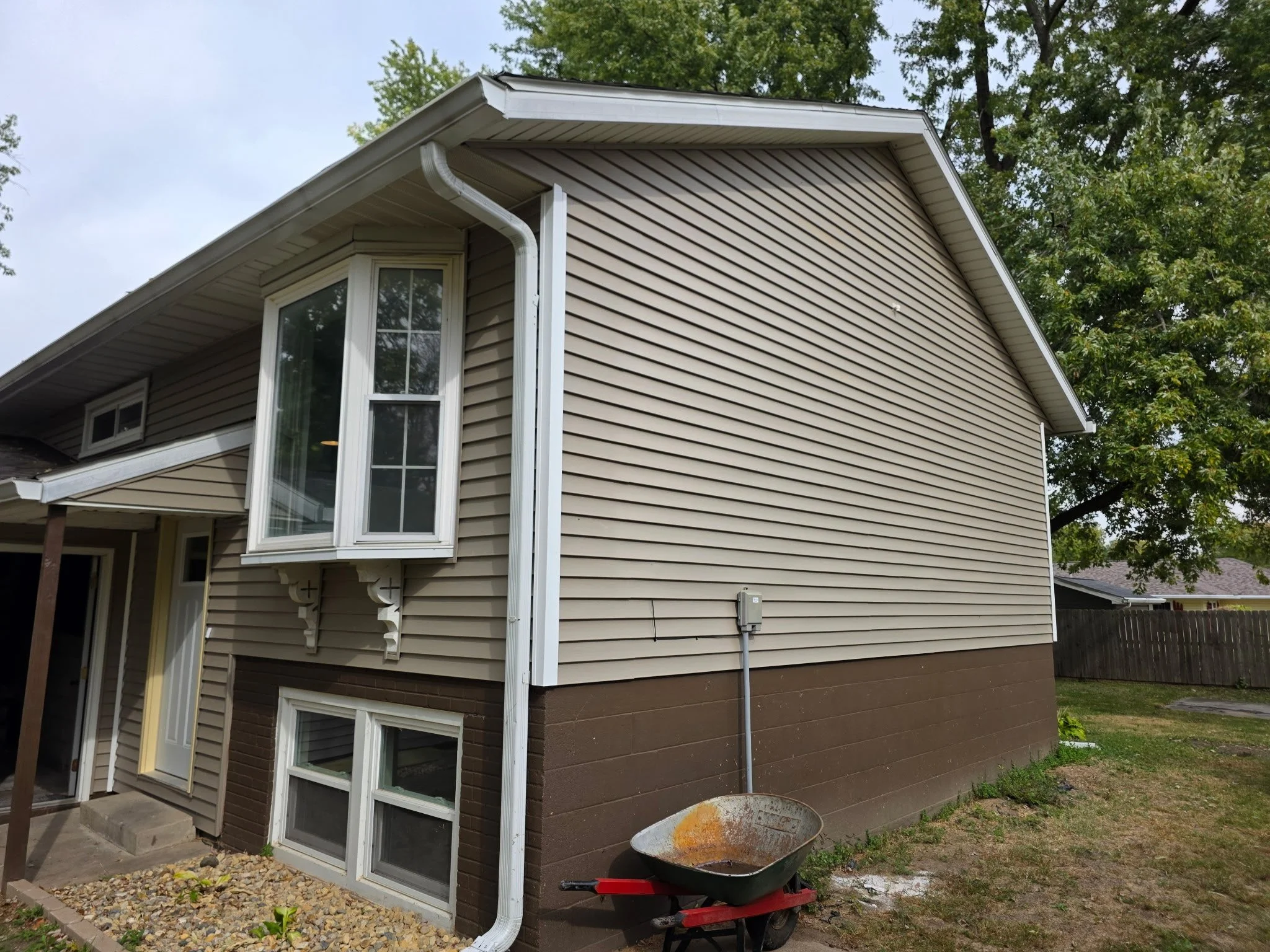 Rear view of a two-story house with beige vinyl siding, brown brick foundation, a wheelbarrow with rust, a gray electrical box, and a large leafy tree in the background.