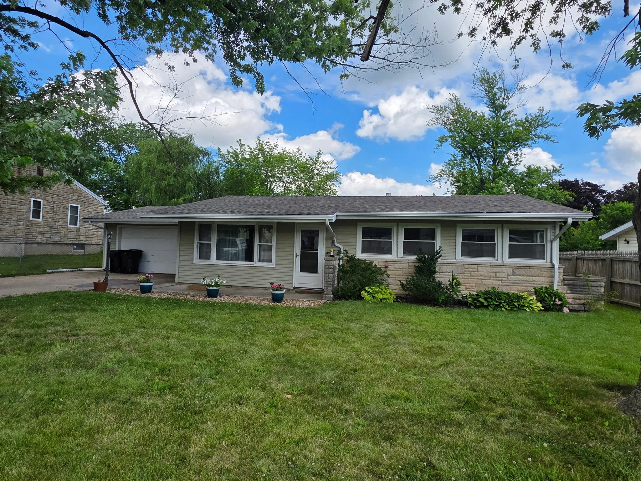 Front view of a suburban house with a lawn, potted plants, and garden bushes under a partly cloudy sky.