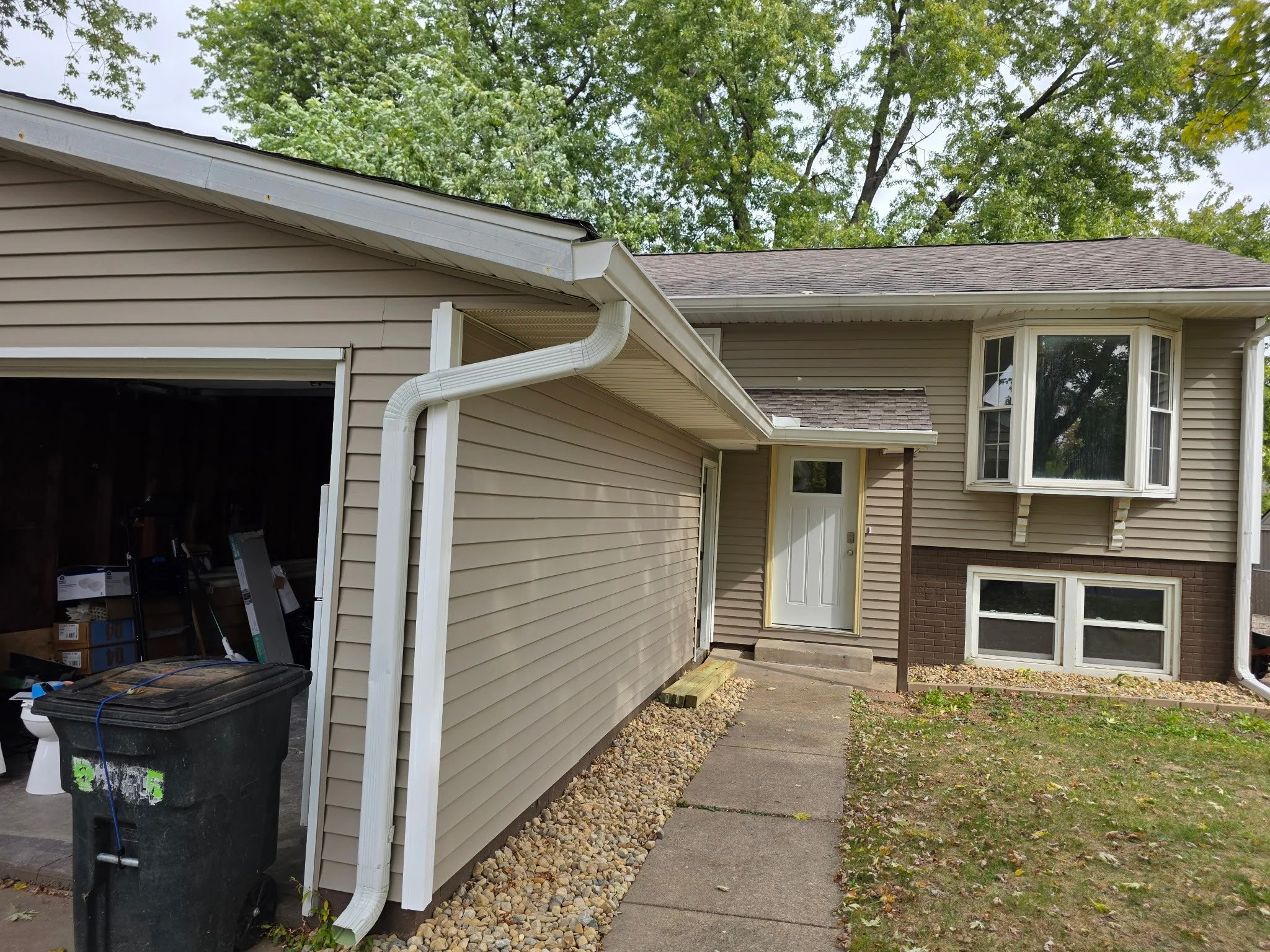 Front view of a beige residential house with white trim, a bay window on the upper level, a small entry porch, and a driveway on the side with a trash bin. The yard has patches of grass and small rocks, and green trees are visible in the background.