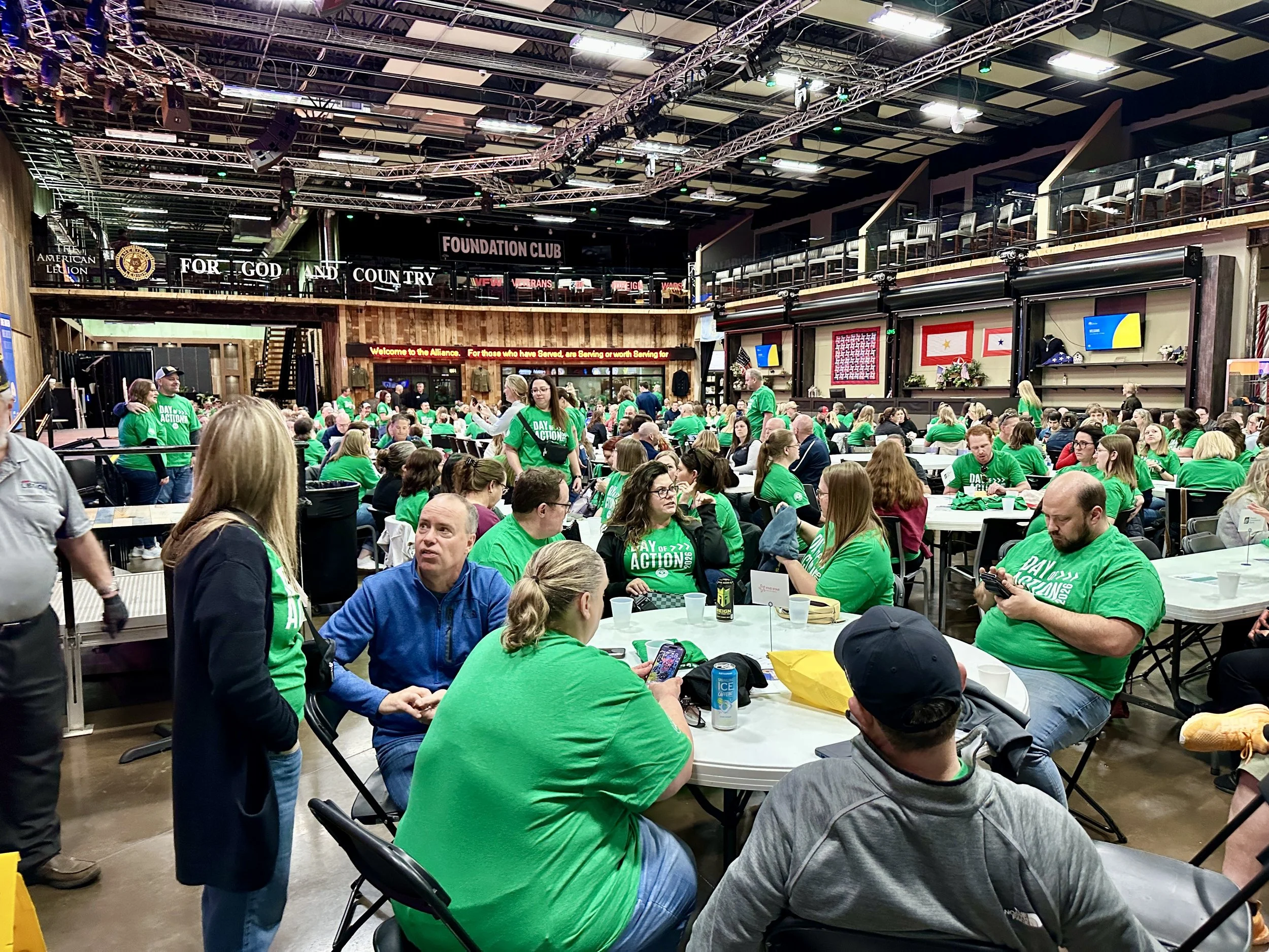 Large indoor event space filled with many people sitting at tables, most wearing green shirts. There are signs on the walls with patriotic and political themes, including flags and the words 'Foundation Club' and 'For God and Country'. The space has a high ceiling with exposed metal beams and a stage area at the back.