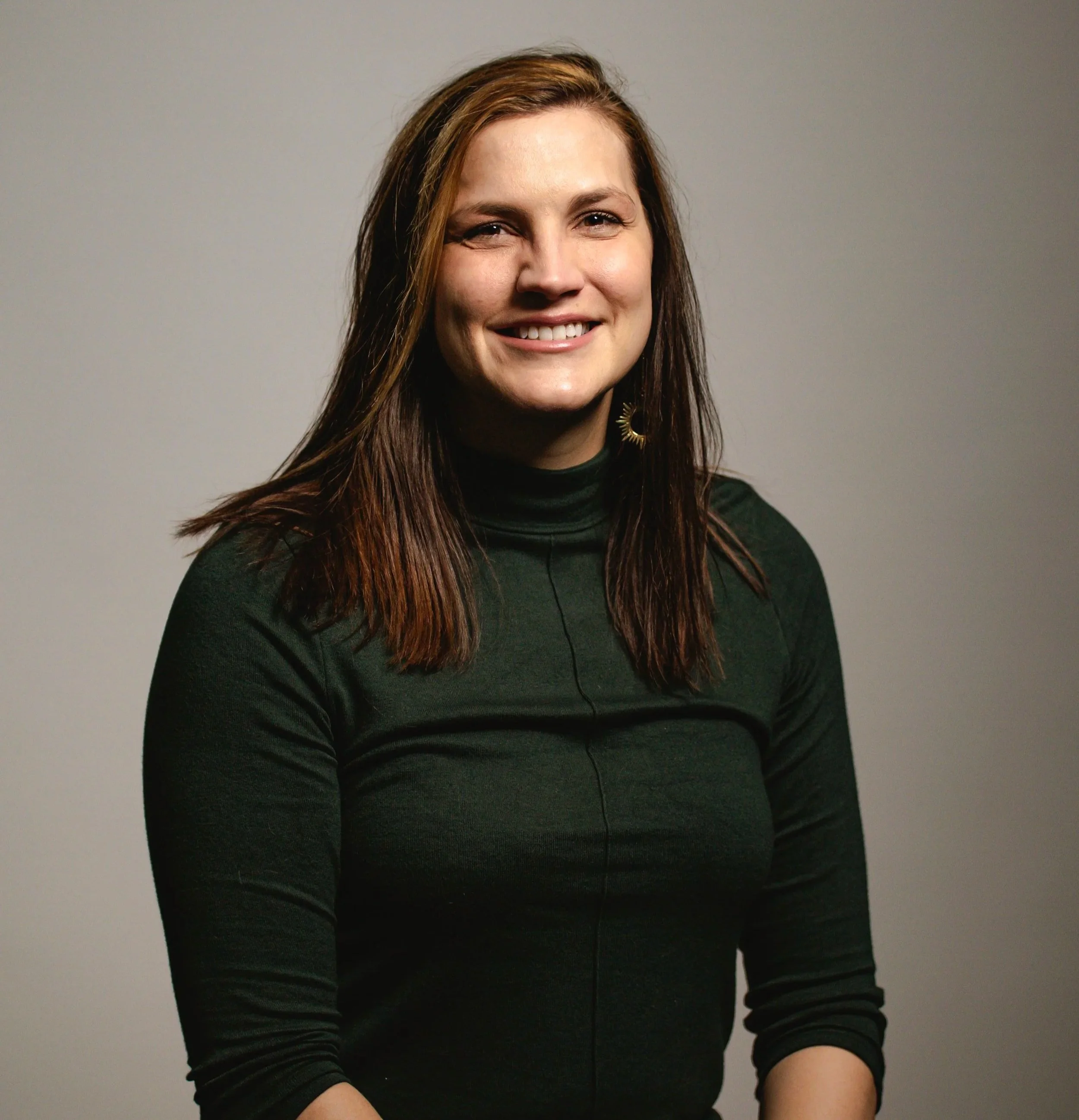 A woman with long brown hair smiling while sitting against a plain gray background.