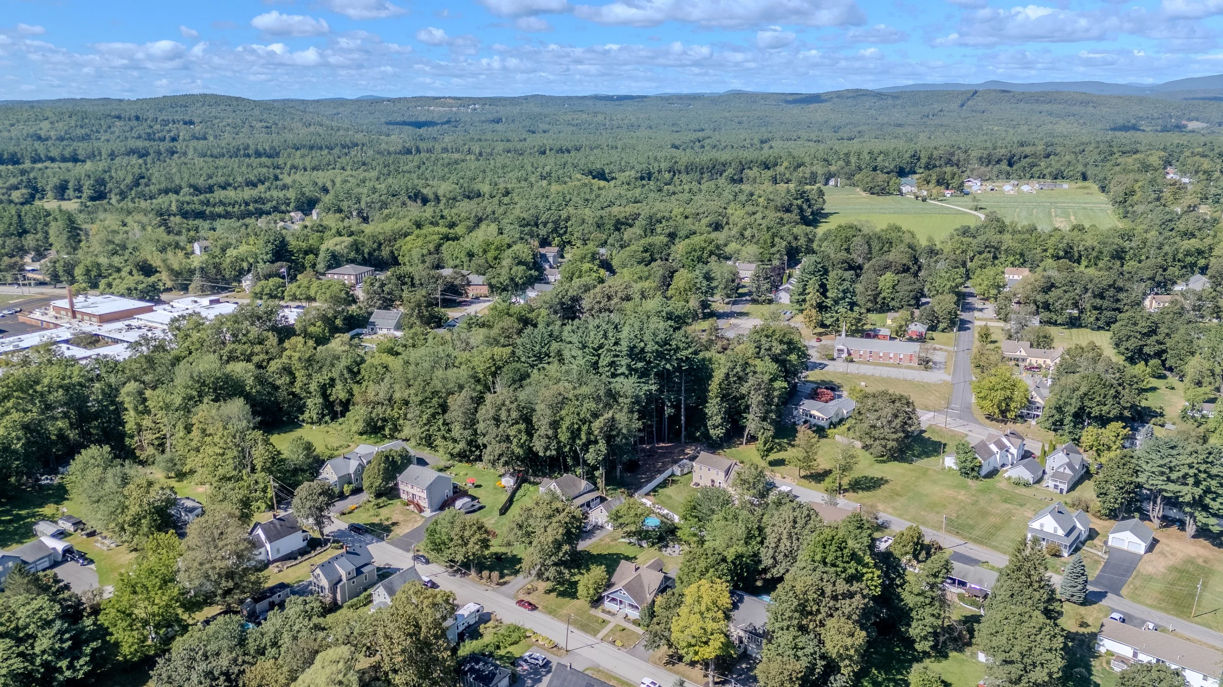 Aerial view of a small town surrounded by green trees and rolling hills, with residential houses, a school, and commercial buildings visible.