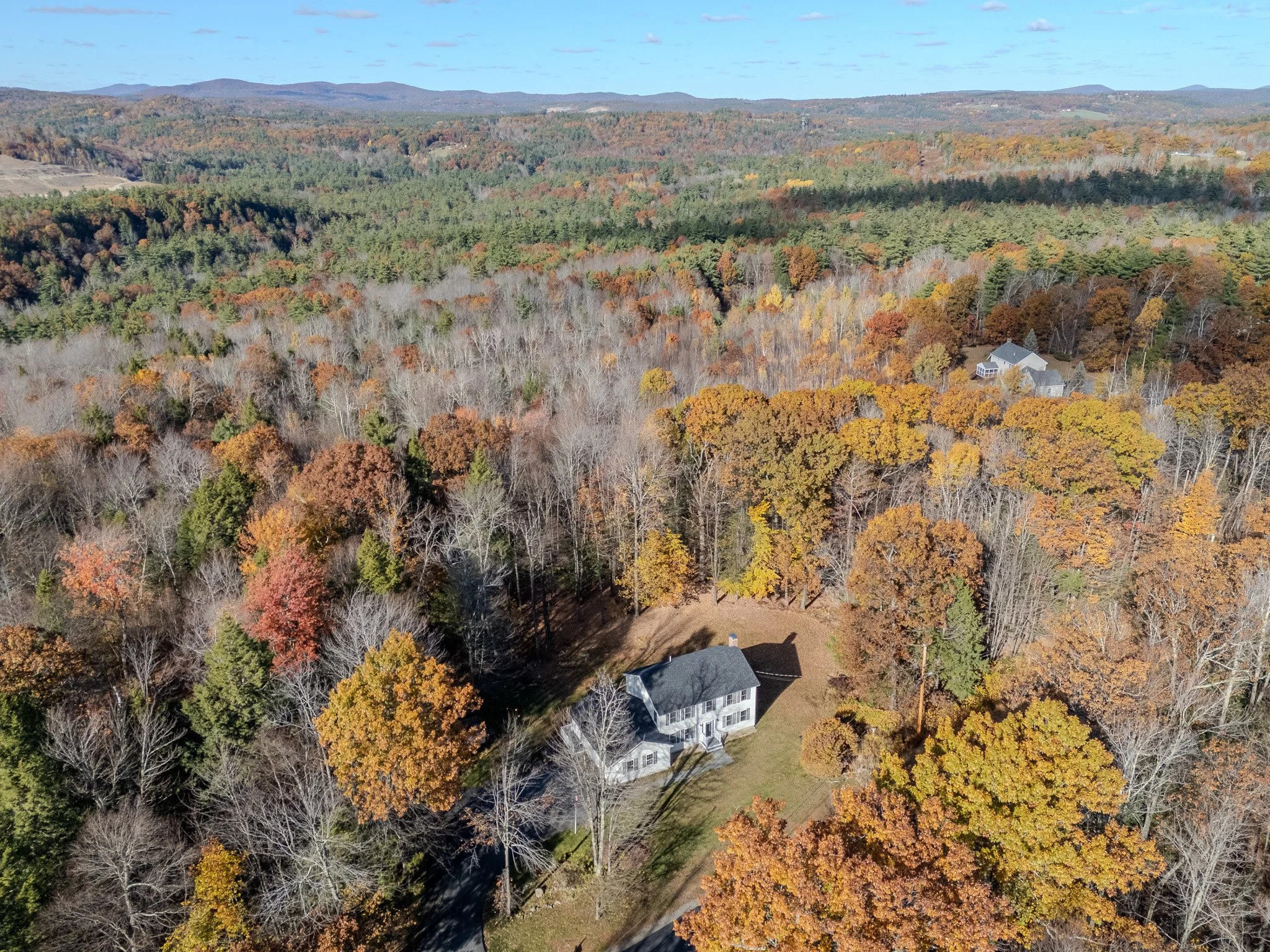 Aerial view of a house surrounded by autumn trees with colorful fall foliage, set in a forested area with distant hills under a partly cloudy sky.